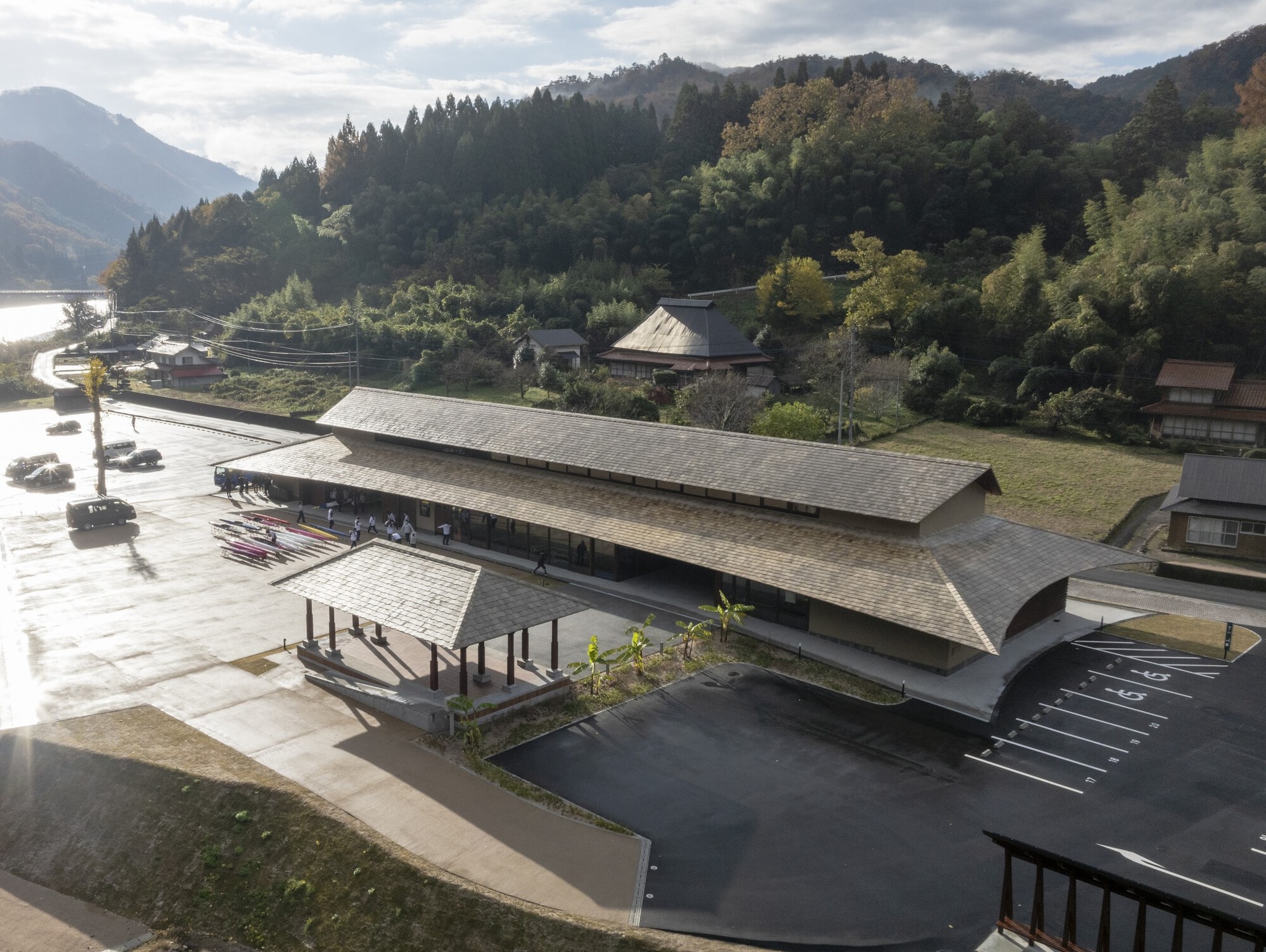 Aerial view of the long hipped roof with shingle cladding, a small pavilion and parking lot below forested hills in autumn