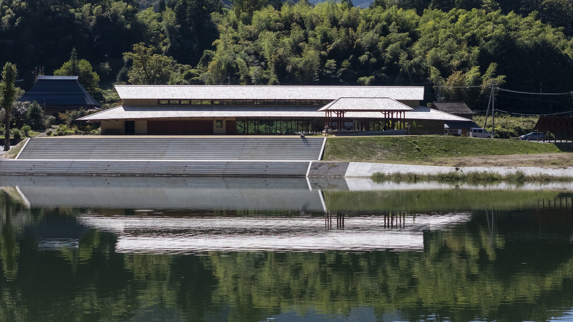 Low pavilion with a shingled roof and stepped stone embankment reflected in still water beneath a wooded slope