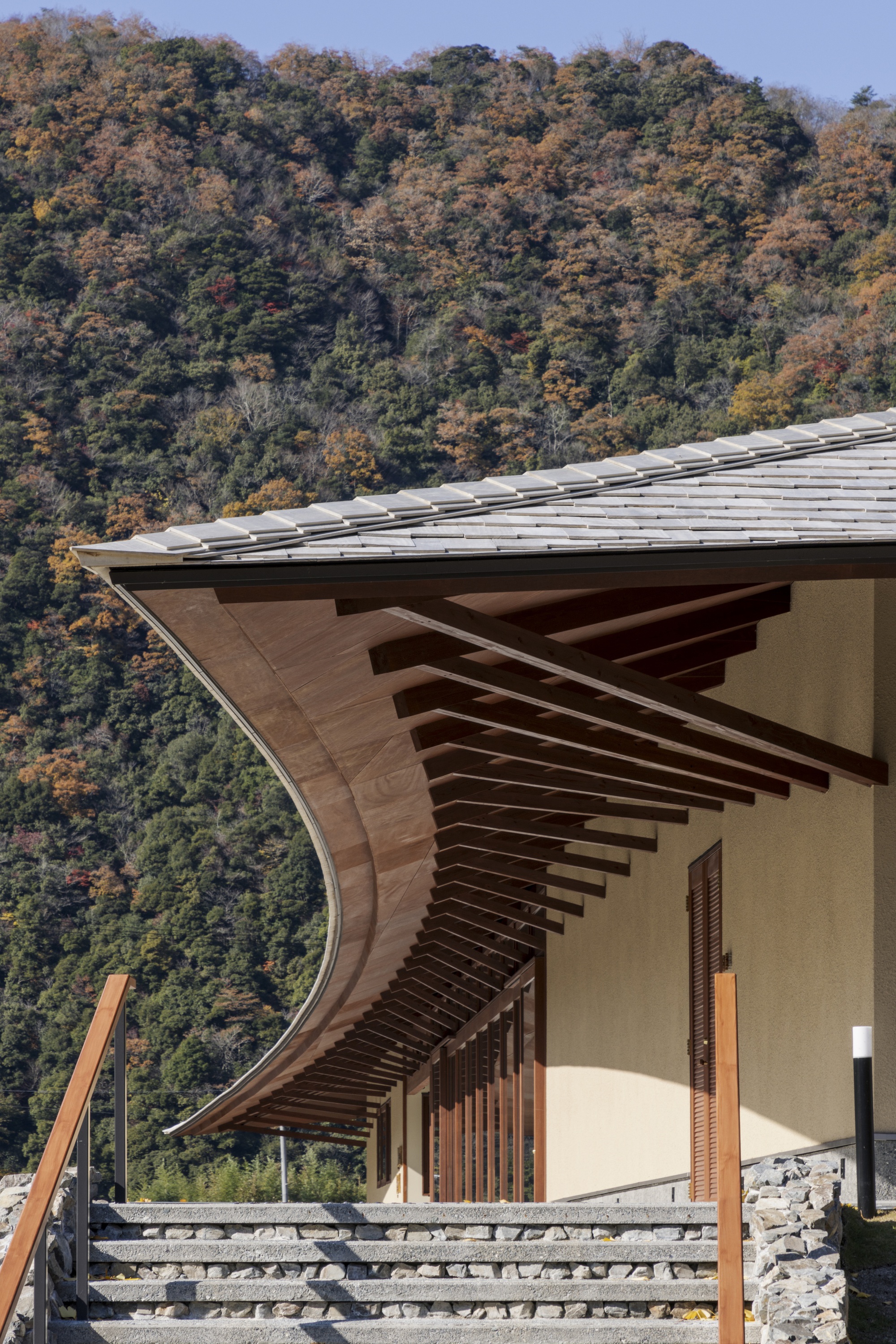 Close view of the curved timber eave with projecting rafters above stone steps, against hills in autumn color