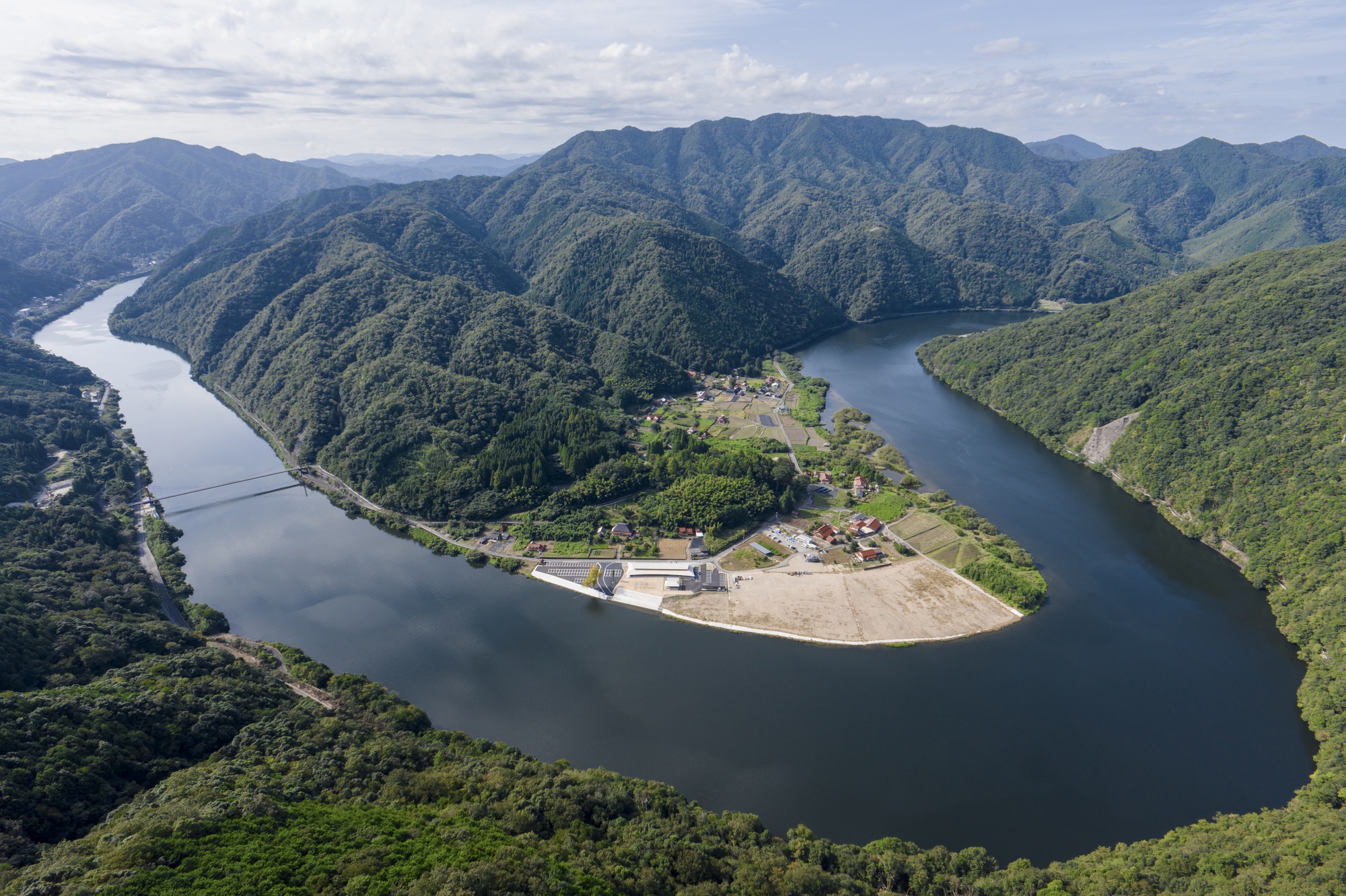 Aerial view of a small settlement on a peninsula inside a river bend, ringed by steep green mountains under scattered clouds