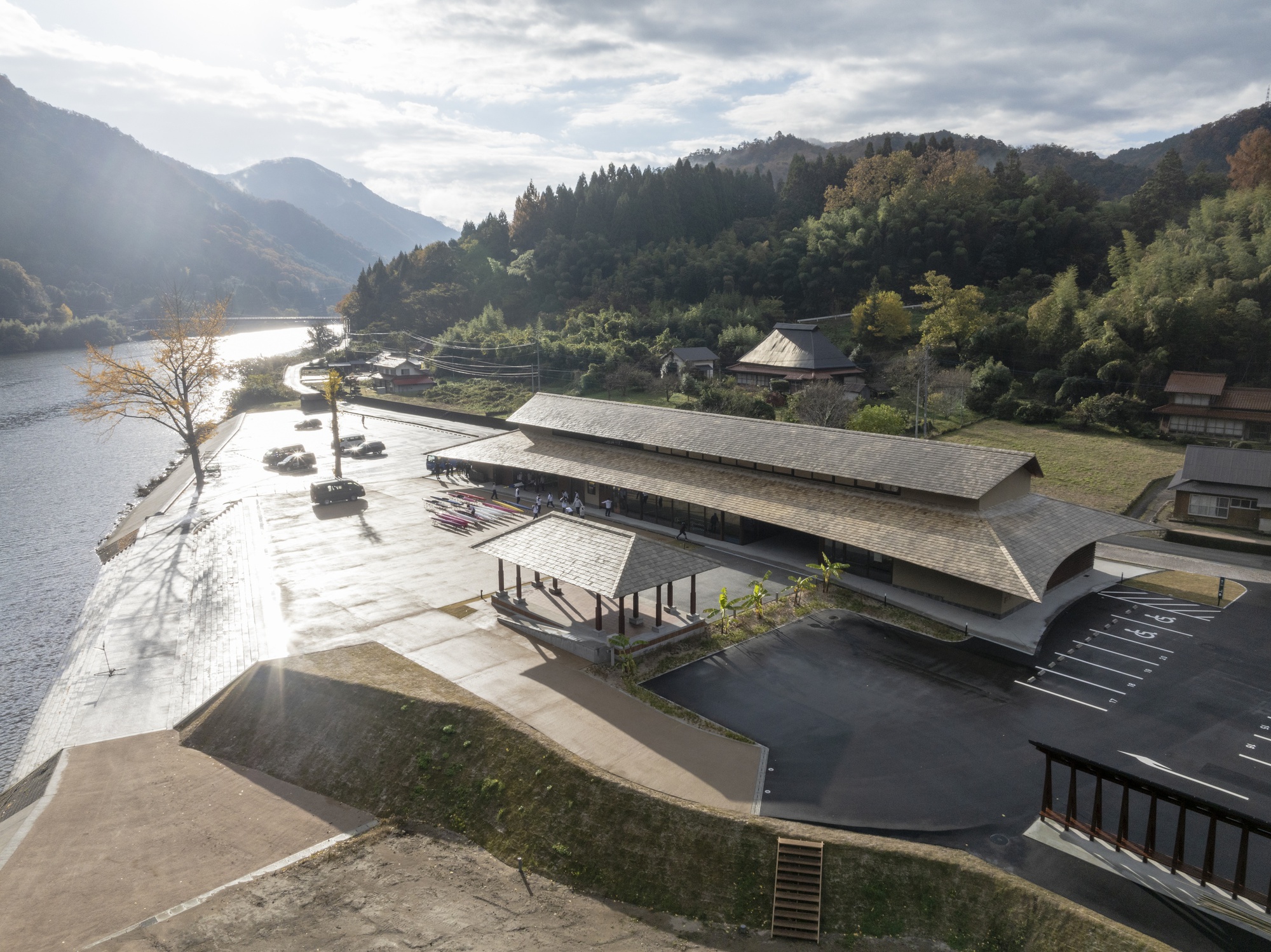 Drone view of the long hipped roof of the lakeside hall beside a paved waterfront plaza in low afternoon sun