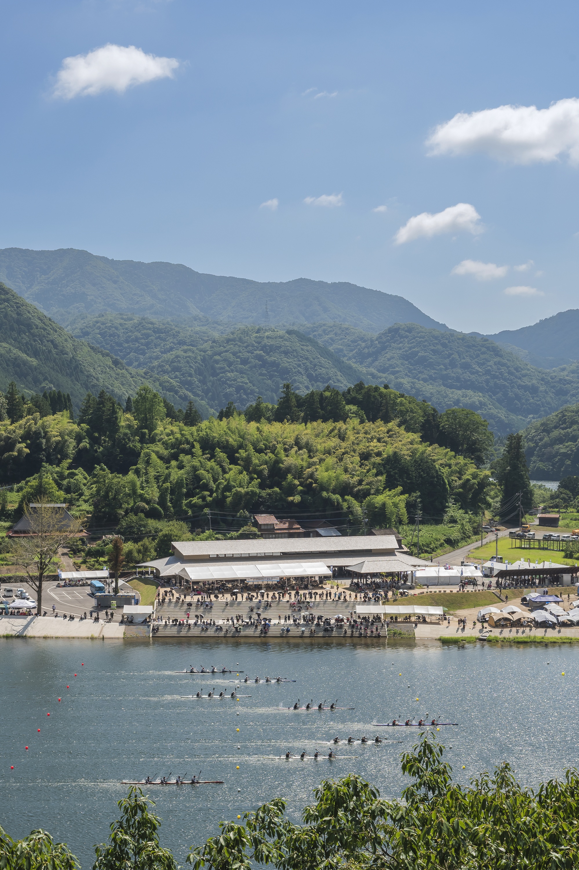 Rowing crews racing across the lake in front of the long-roofed building and crowded embankment on a clear summer day