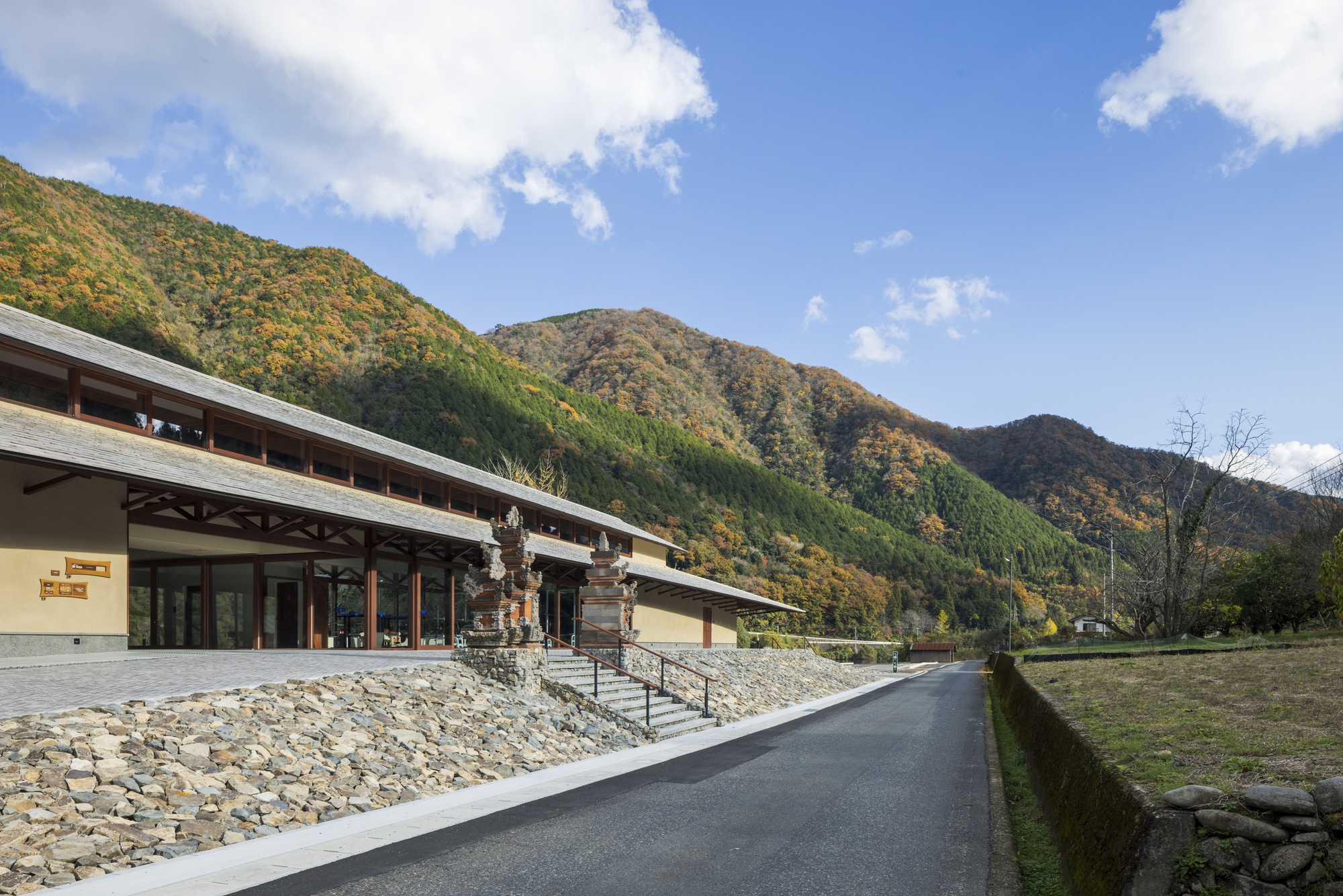 Side view of the tiled-roof pavilion with carved stone gate figures, a rubble stone stair and autumn hills behind