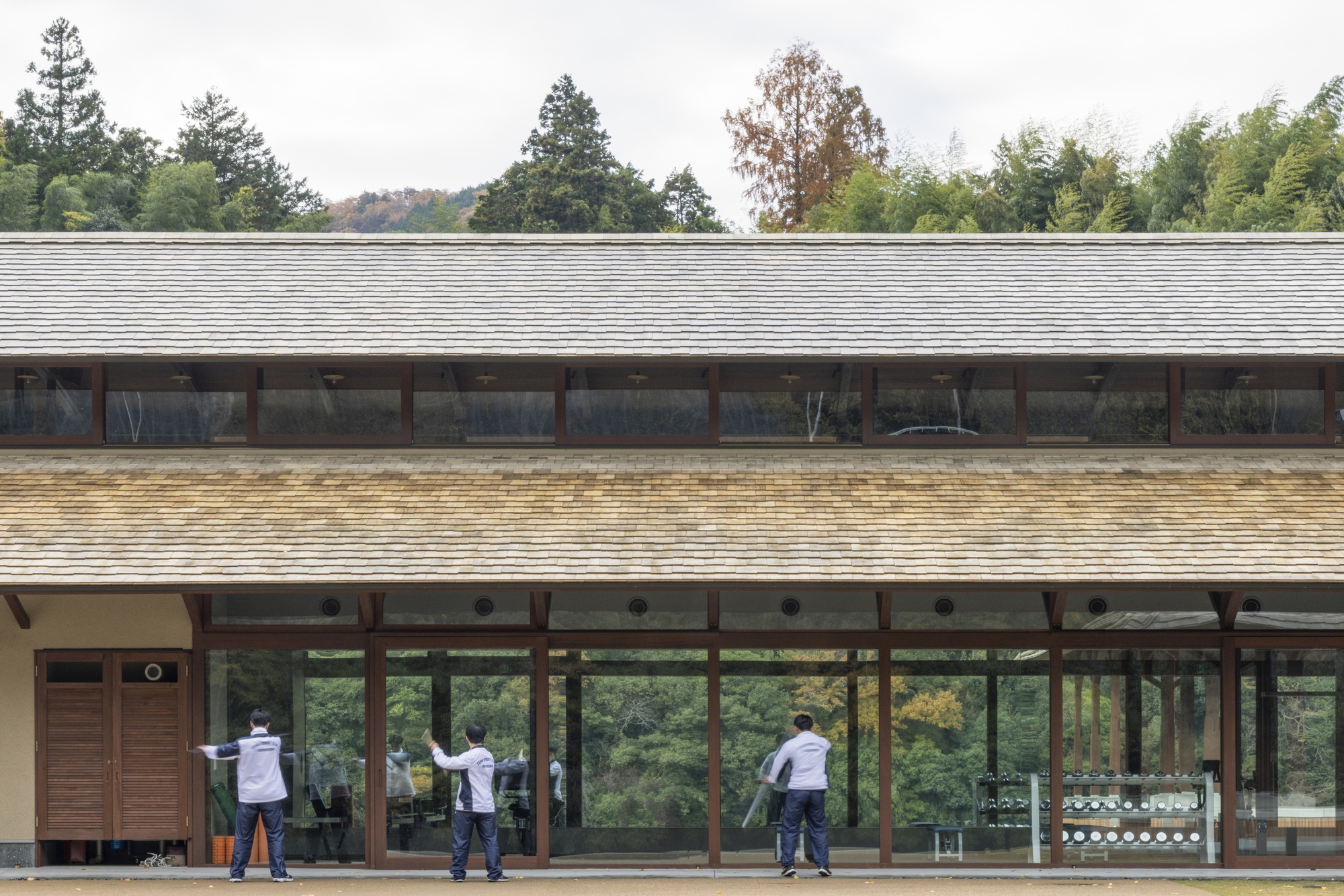Three people practicing tai chi in front of the glazed timber facade with its layered shingle roofs and surrounding trees