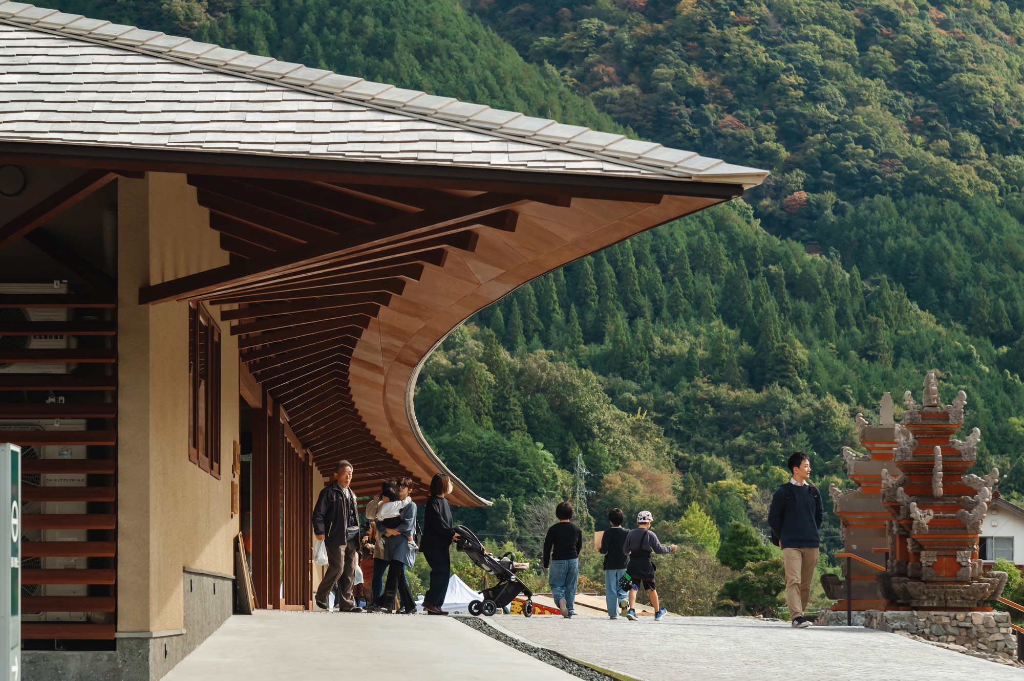Curved timber eave sweeping over a walkway where visitors and a stroller pass, with forested hills and a carved red stone pagoda behind