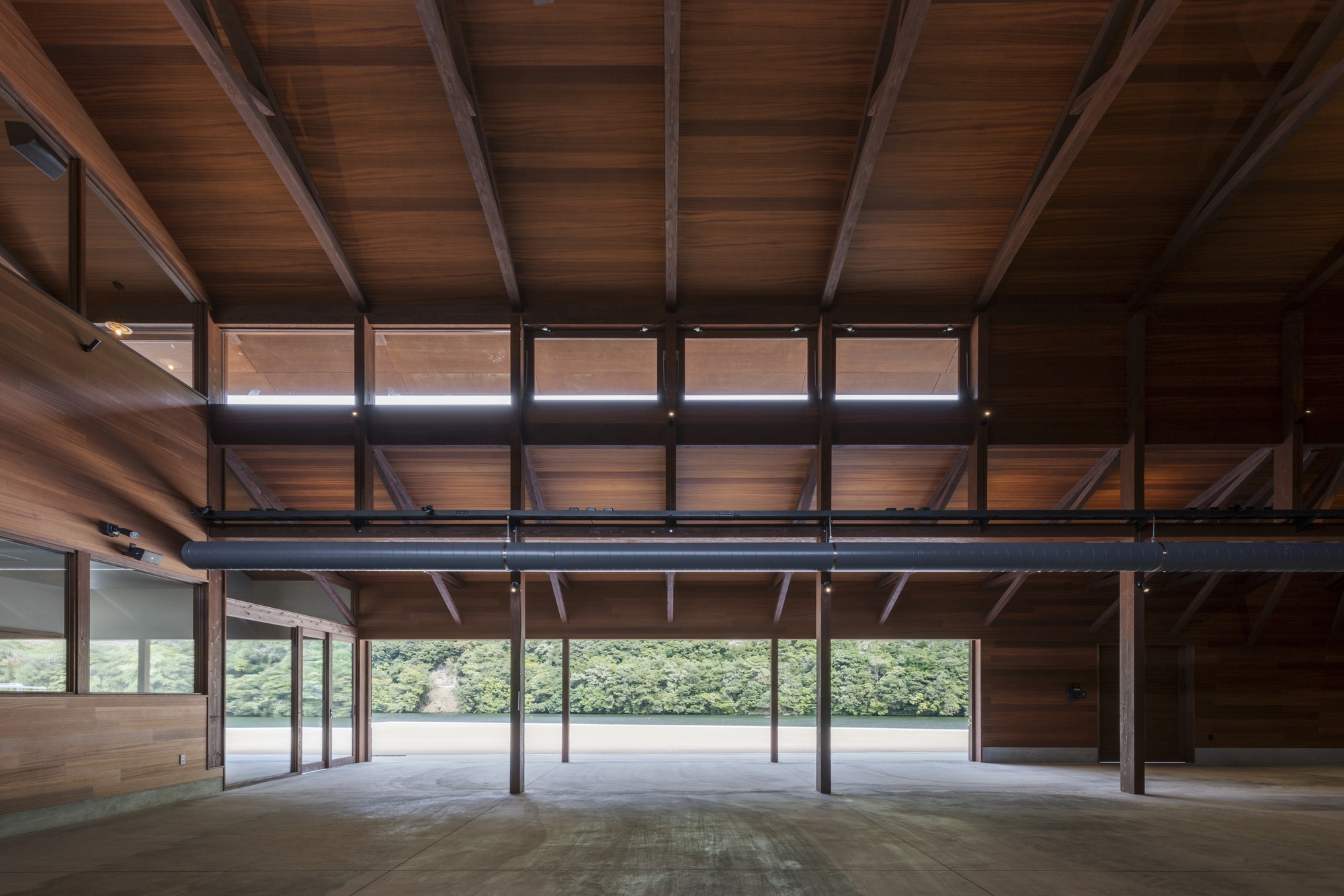 Interior of a timber-lined hall with exposed rafters and clerestory windows, opening to a green hillside through a glazed wall