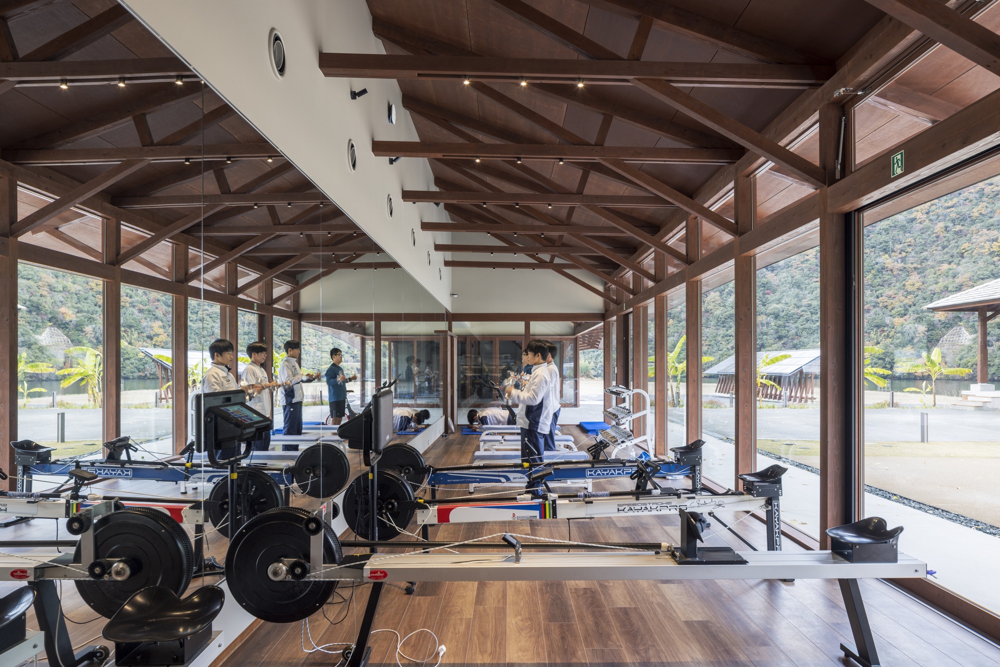 Rowing machines in a timber-framed training room with floor-to-ceiling glass, as athletes stand in line against a hillside view