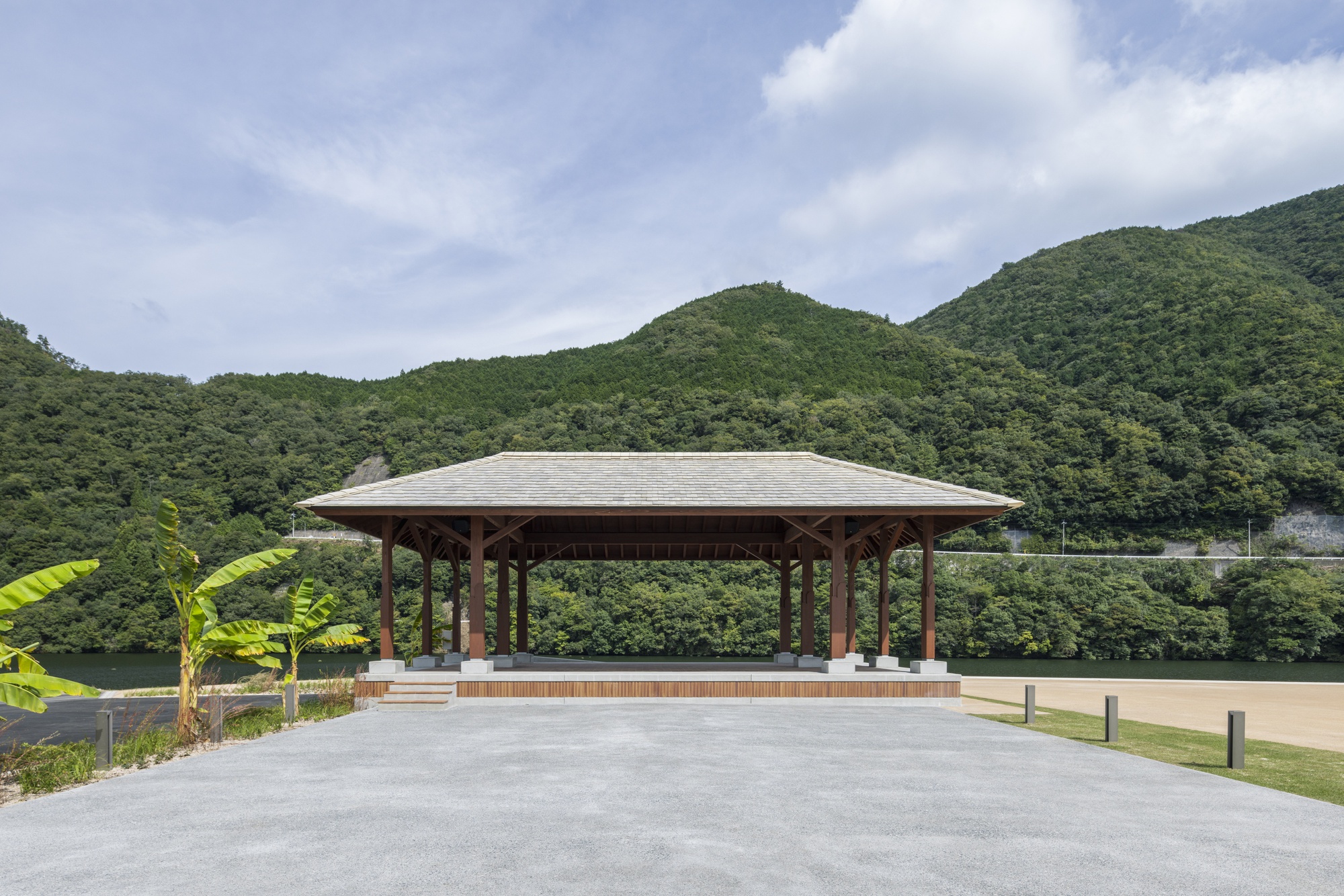Open timber pavilion with a hipped tile roof on a raised platform, set before forested hills under a cloudy sky