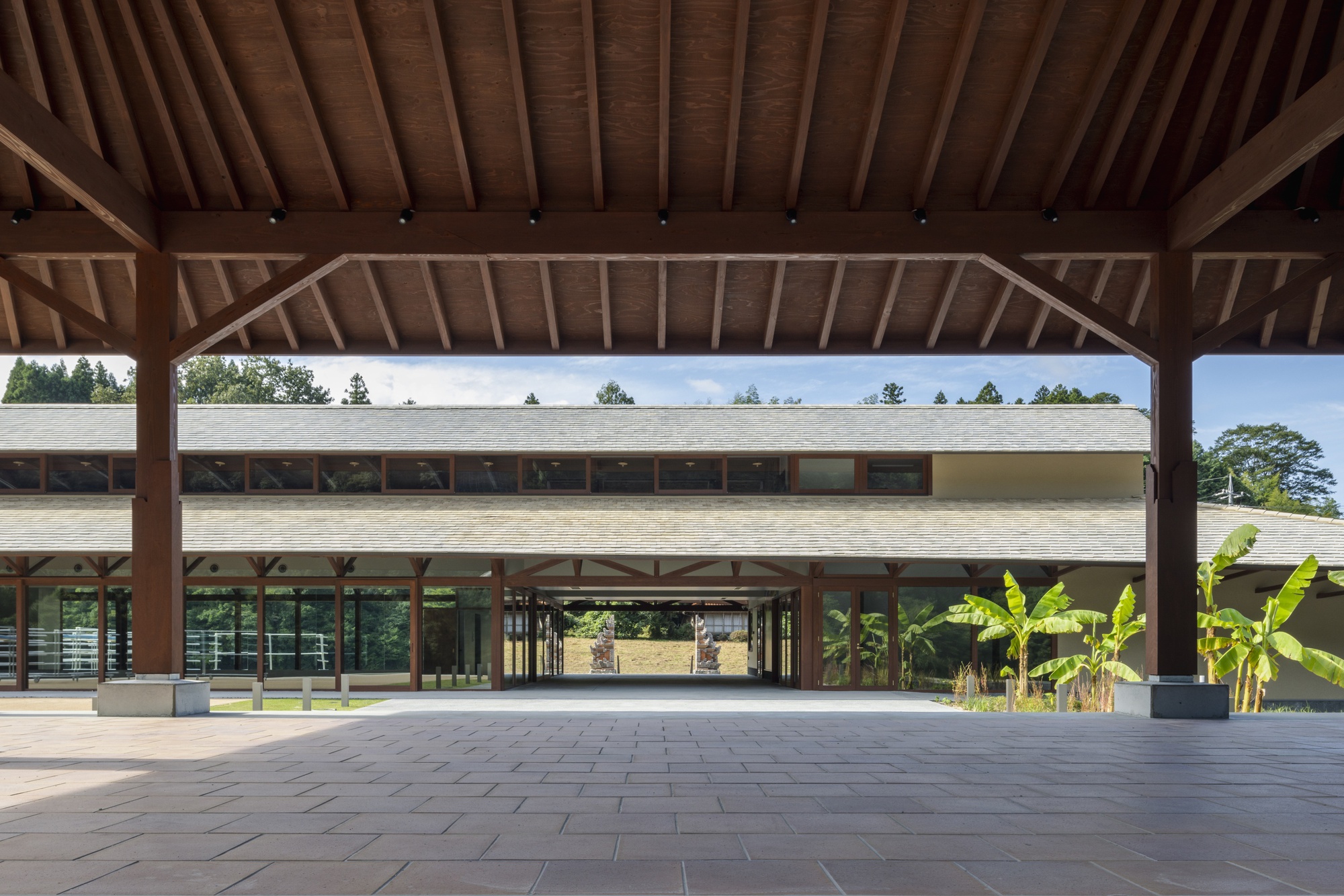 Open timber-framed porch with heavy columns and exposed rafters facing a low slate-roofed hall, banana plants at the right under a clear sky