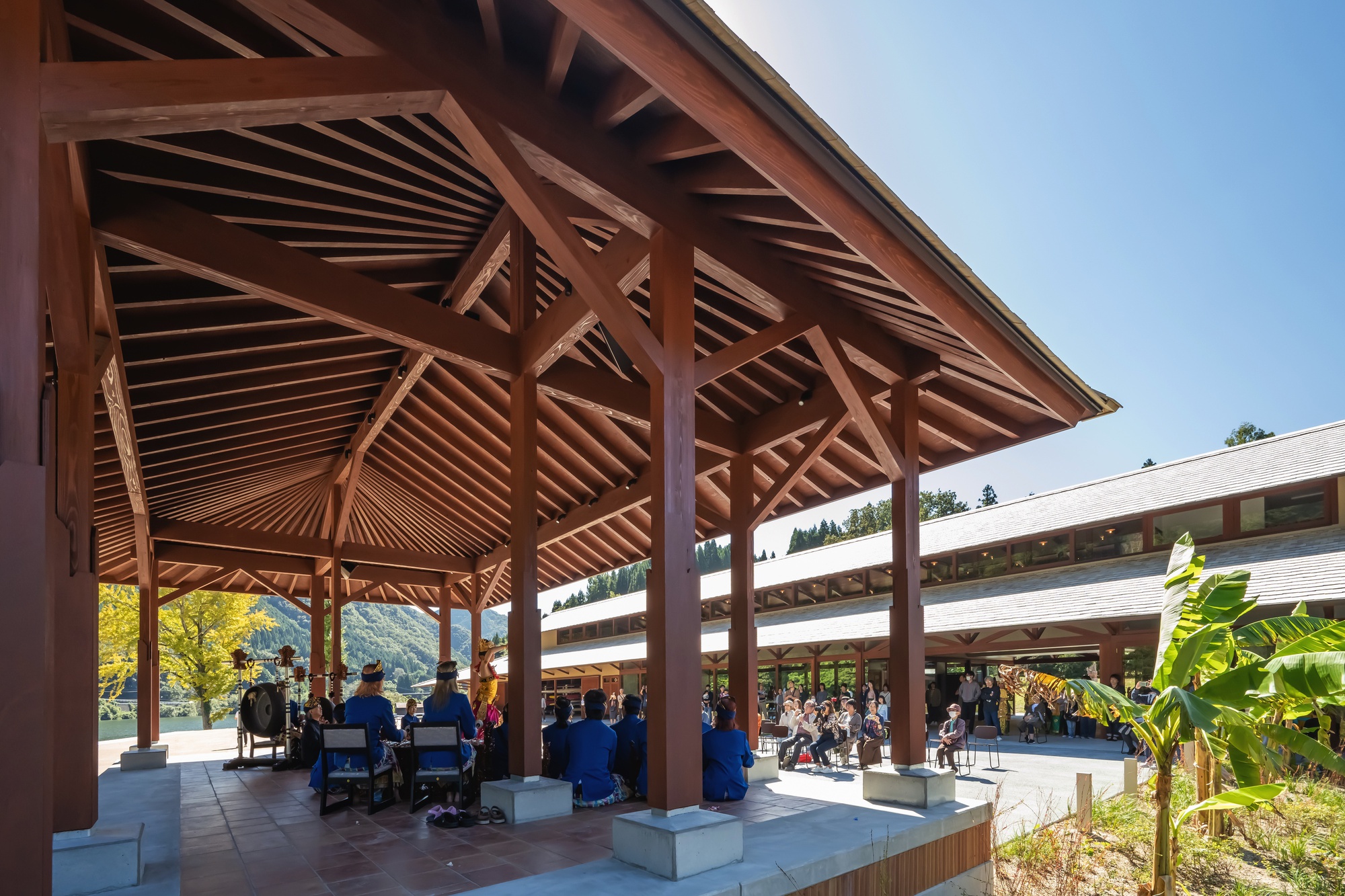 Gathering of seated people and a drum under a wide timber pavilion roof, long slate-roofed hall beyond in autumn sunlight