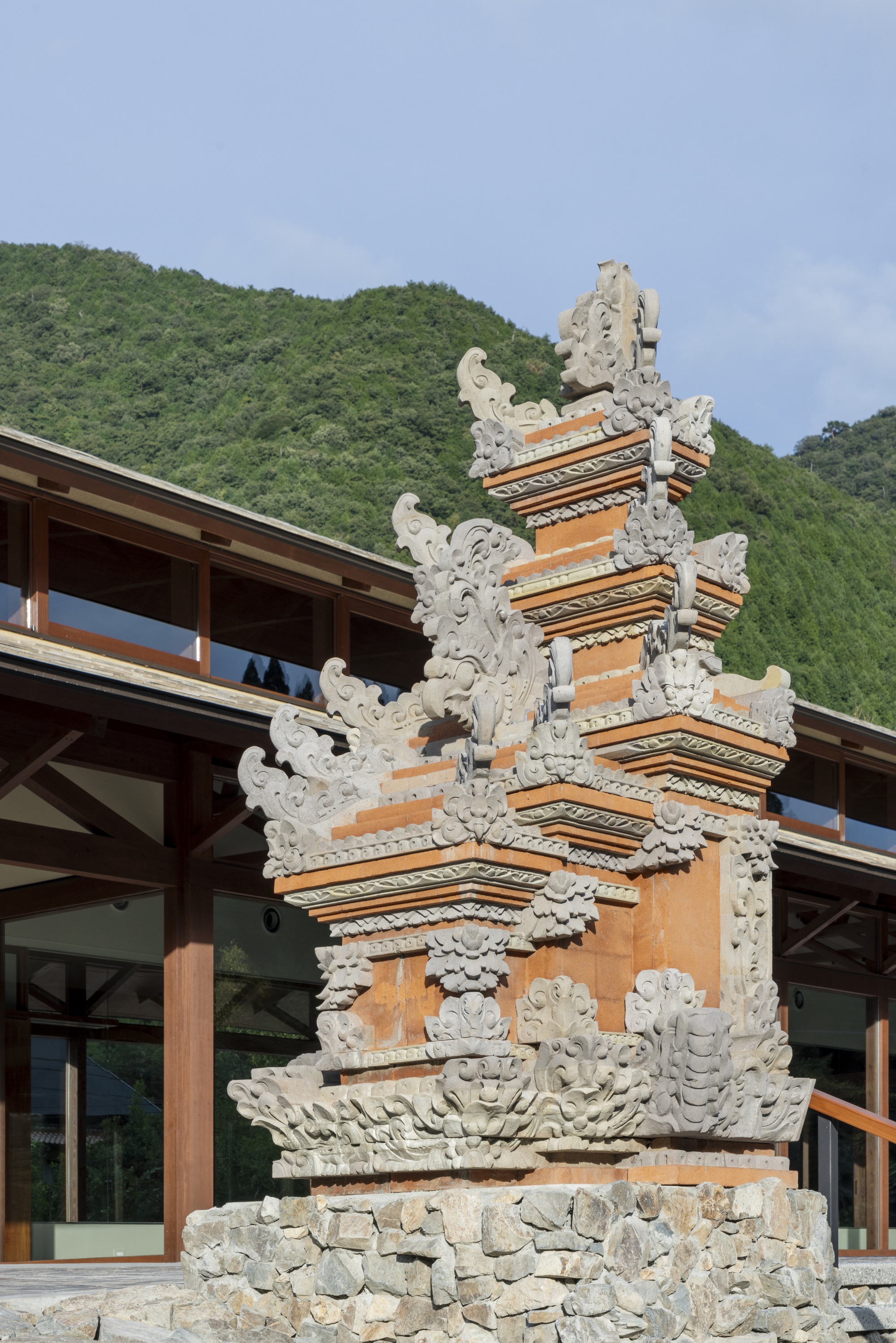 Carved stone and terracotta shrine-like gate with ornate scrollwork on a rubble base, wooded green hillside rising behind