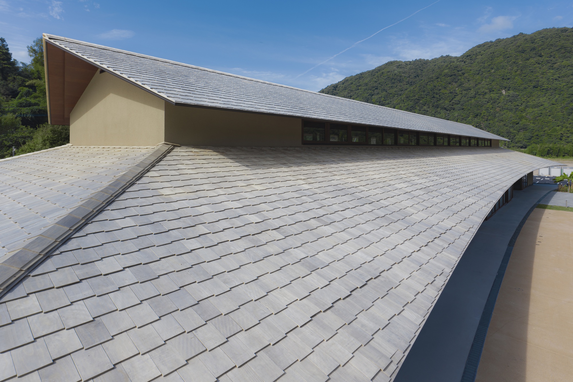 Curving slate-tiled roof sweeping along the hall with a clerestory band of windows, forested hill and blue sky behind