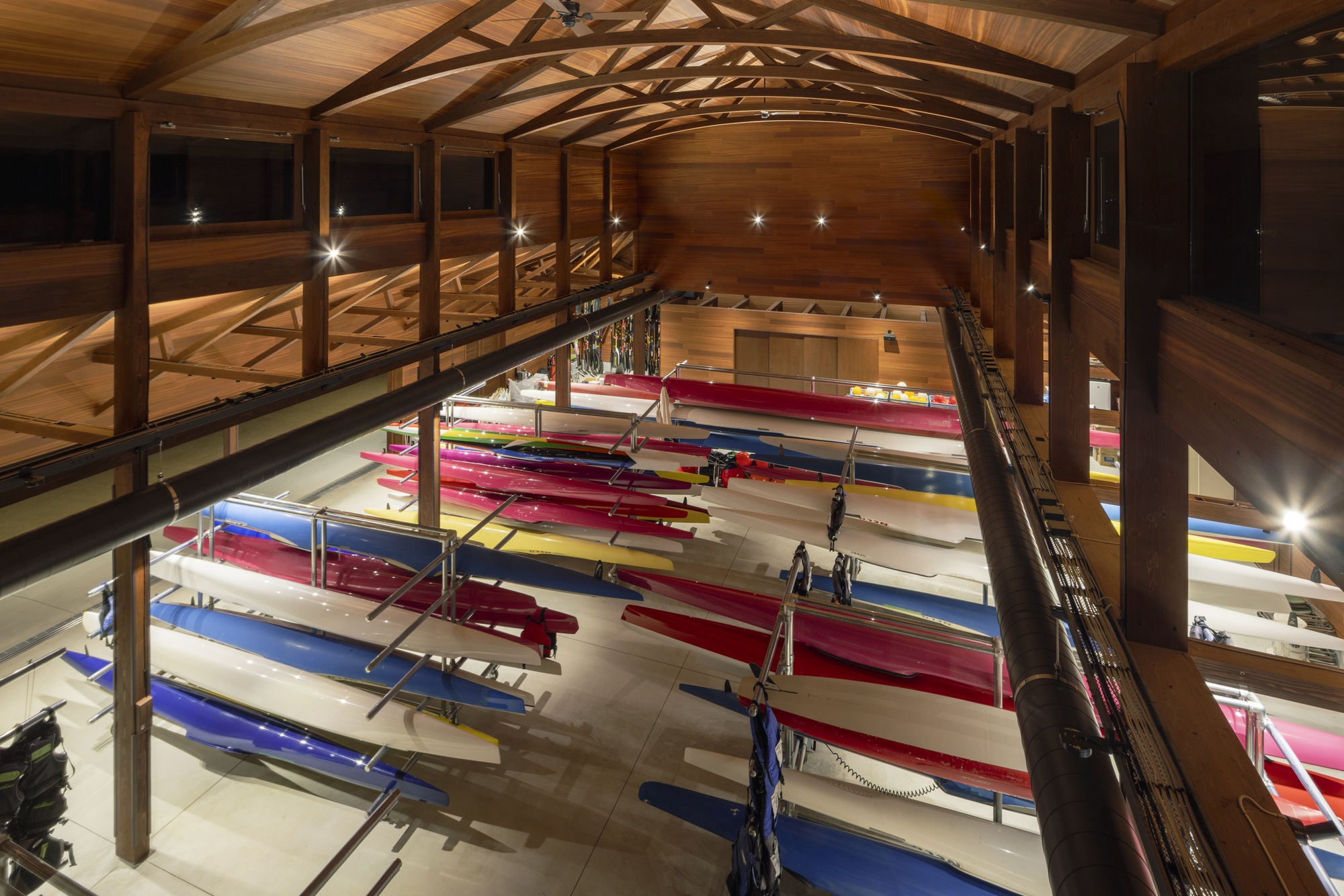 High view into the timber boathouse hall with racks of red, blue and white rowing boats under arched wooden roof trusses at night