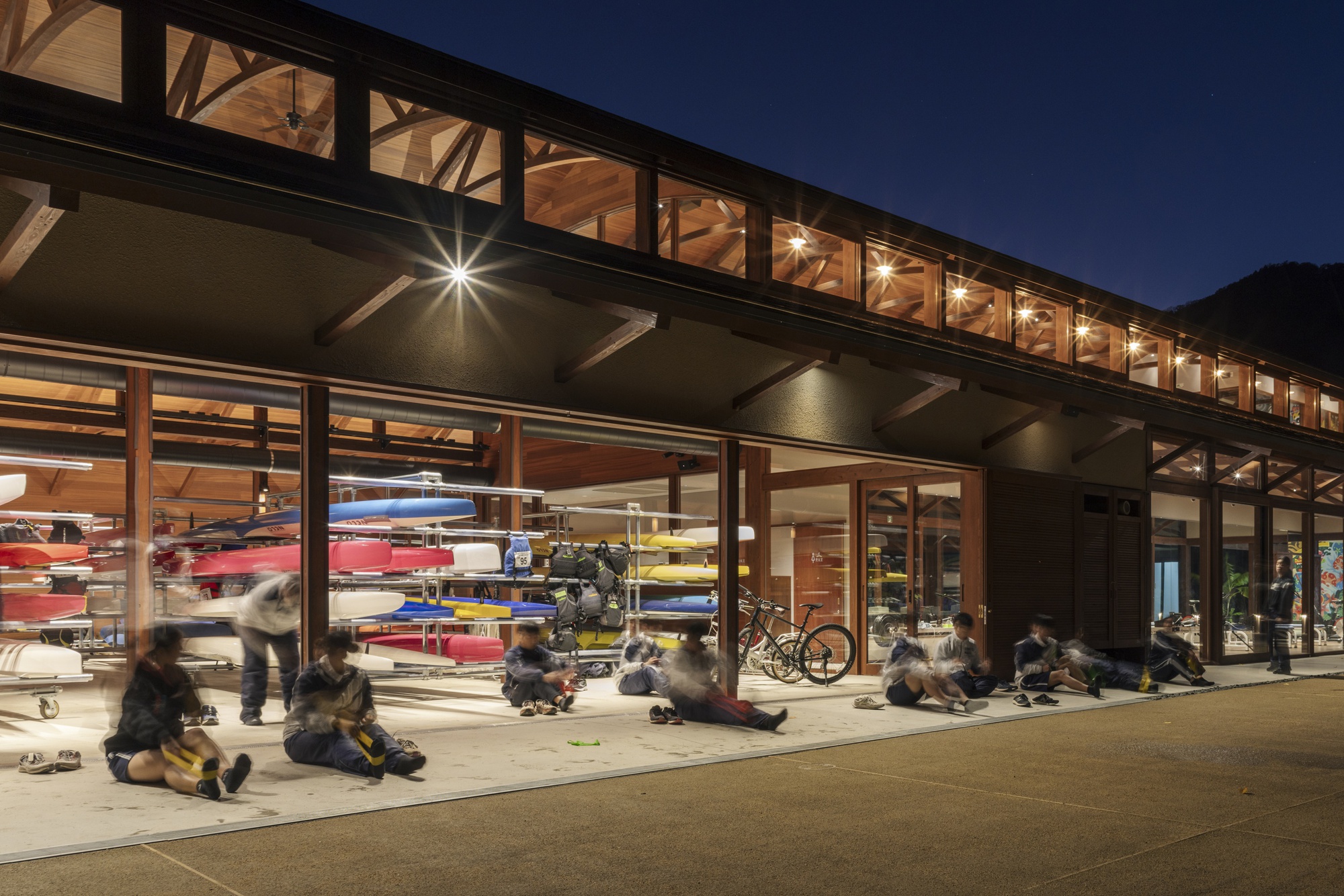 Boathouse at dusk with glass bays open to stacked boats and a bicycle, seated people blurred in the foreground and lit clerestory above