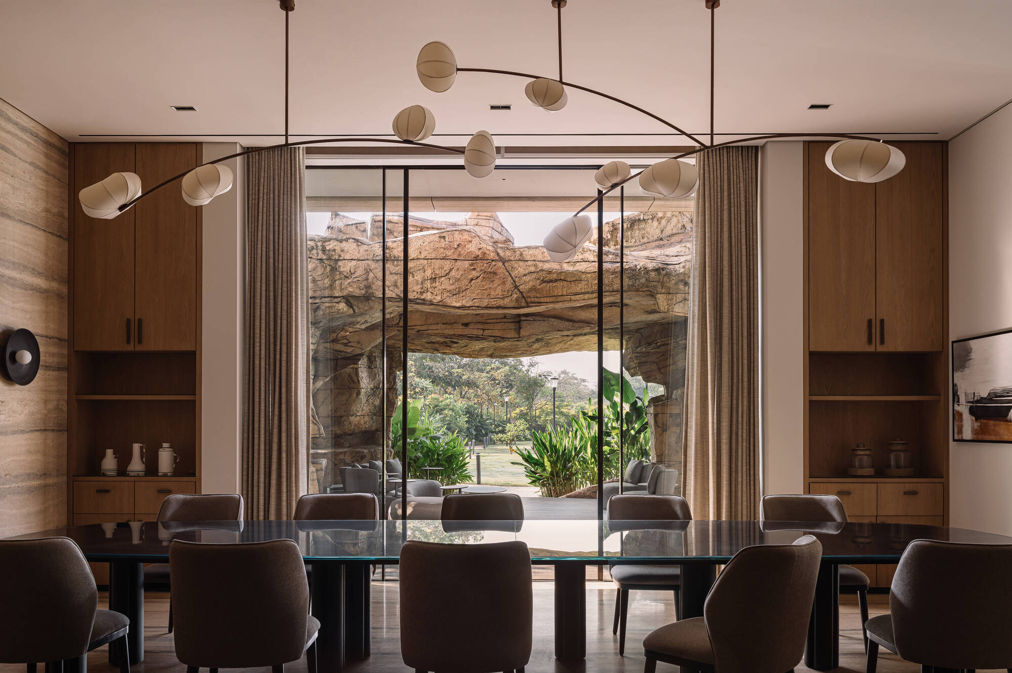 Dining room with a long glass table and branching pendant lamp facing a rock formation through floor-to-ceiling glass