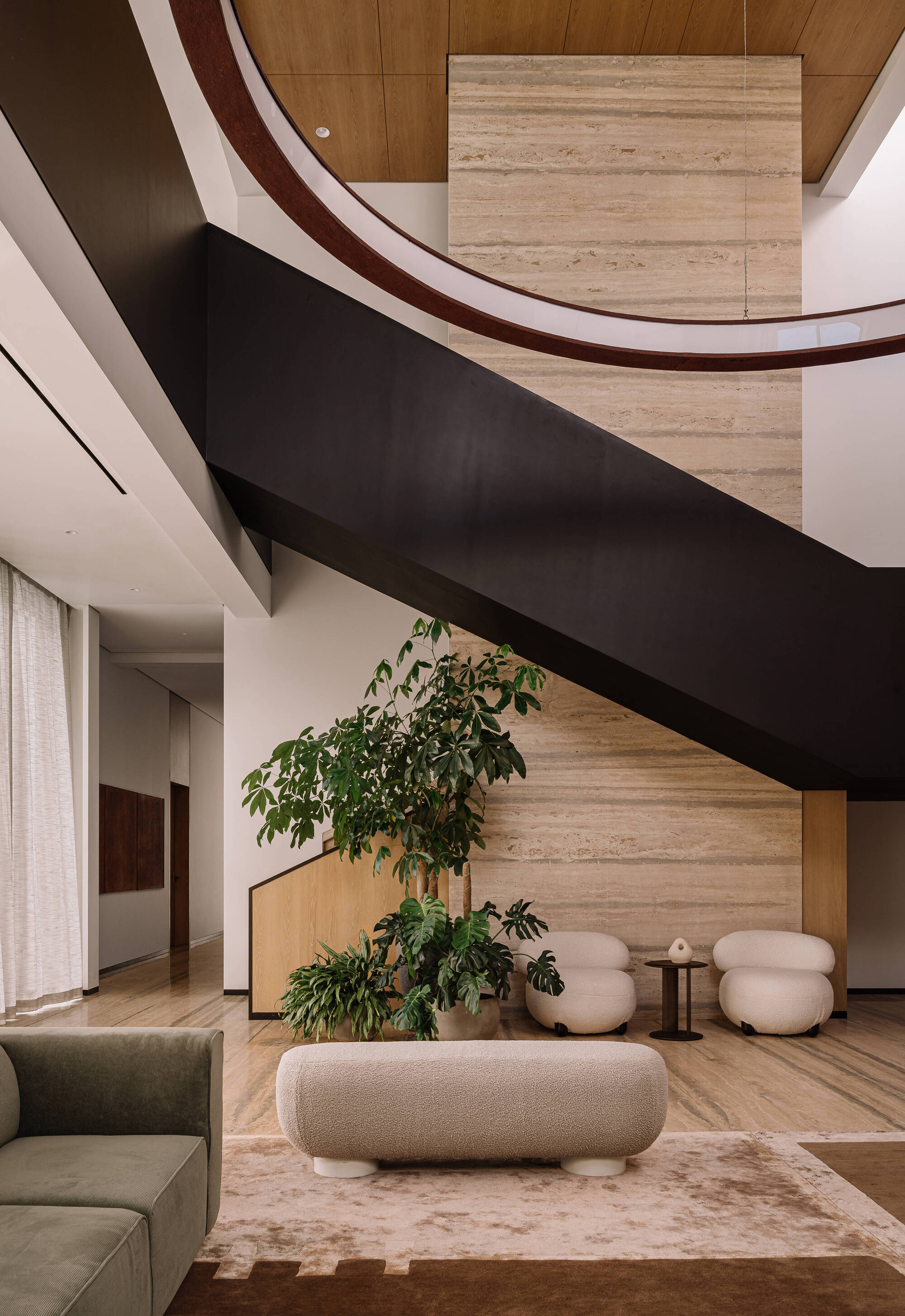 Double-height lobby with a dark floating staircase, travertine wall and a potted tree beside low boucle seating