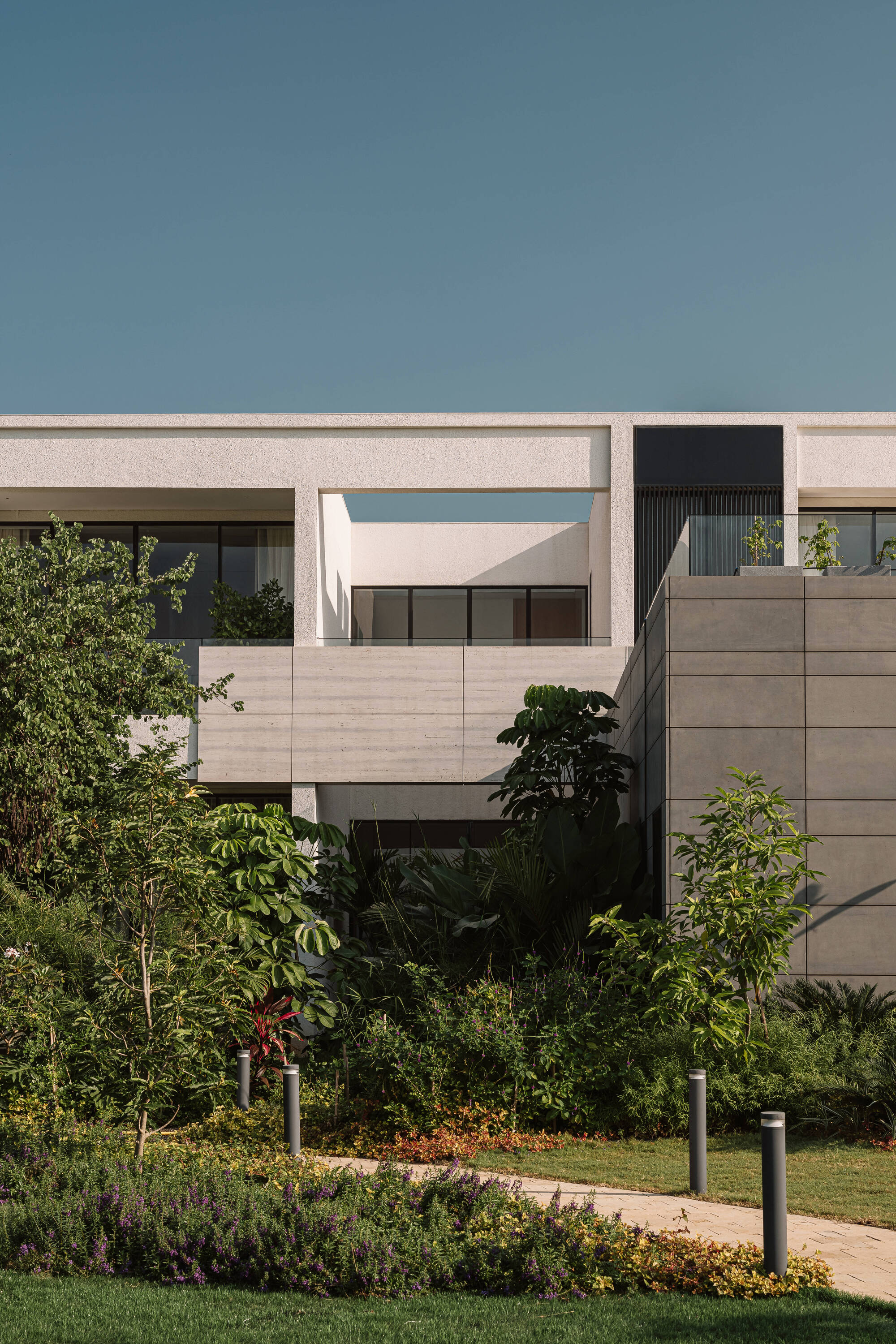 Stepped concrete and stone facade behind layered planting and a paved path with bollard lights under a clear sky