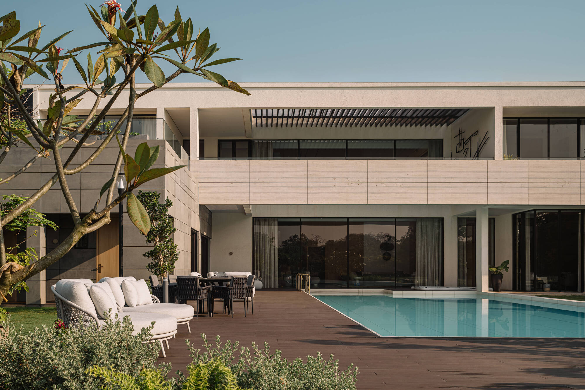 Two-storey stone-clad house facade above a timber pool deck, with white lounge chairs and a frangipani branch in warm evening light