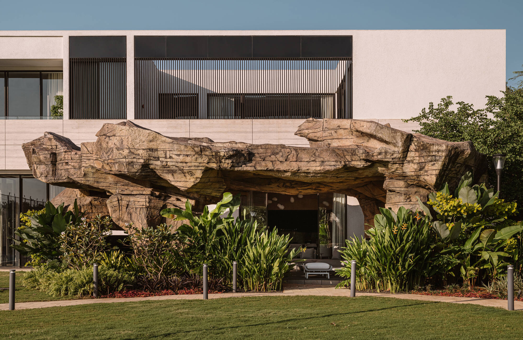 Rock-formed canopy sheltering a lounge beneath the upper volume, ringed by banana-leaf plants and a trimmed lawn in low sun