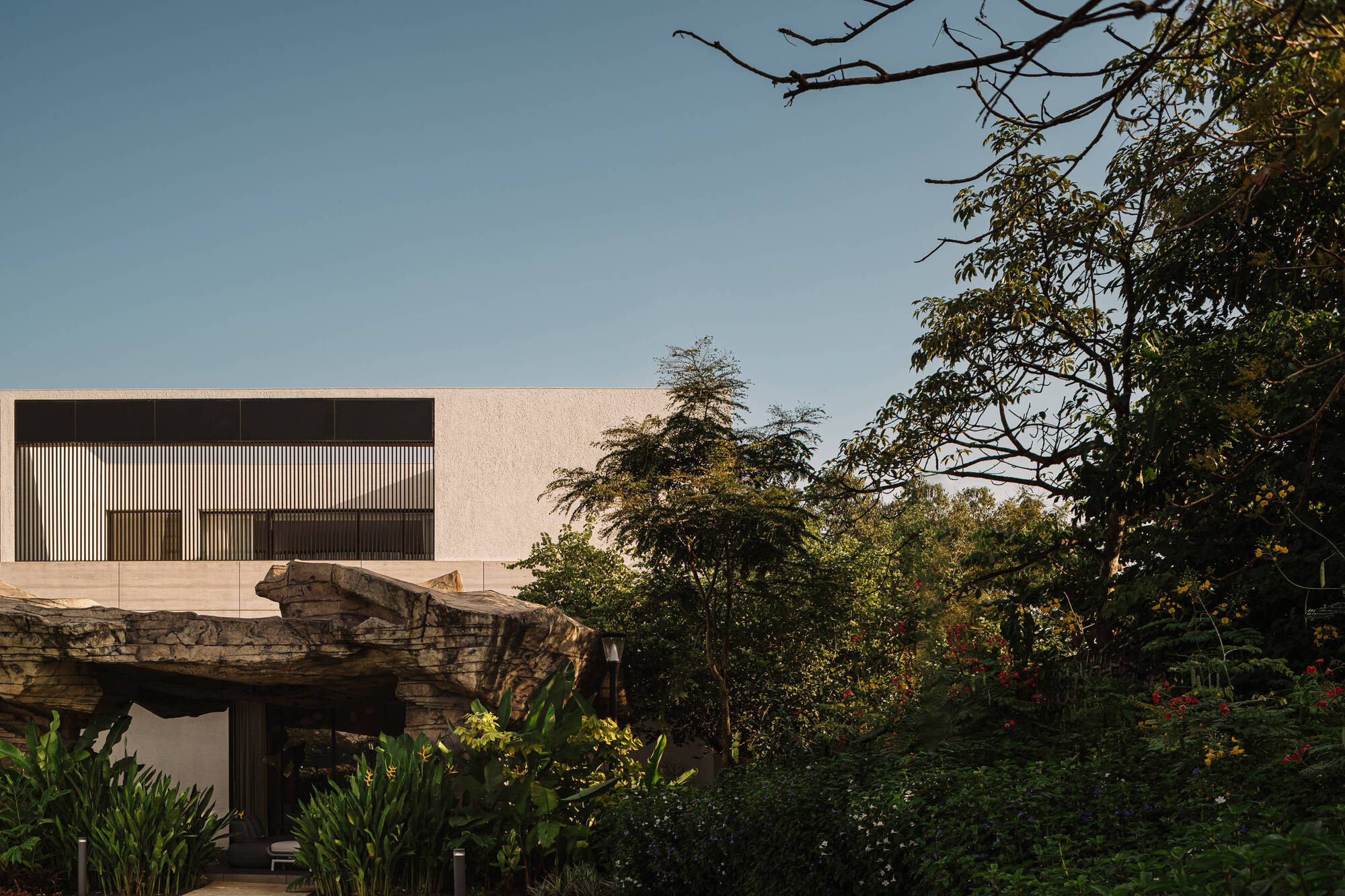 Slatted upper-floor facade rising behind the rock canopy, framed by dense trees and red flowering shrubs under a clear dusk sky
