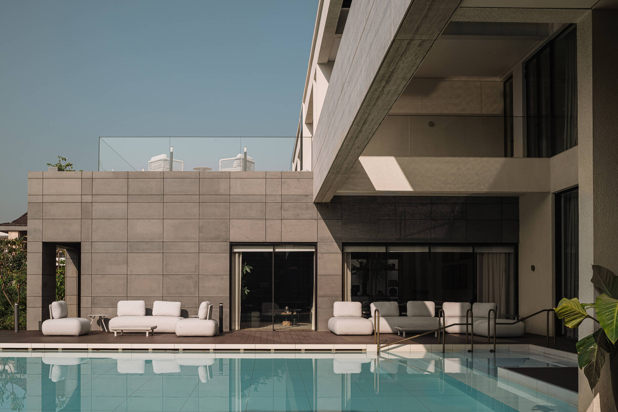 Poolside terrace with white modular sofas against a gridded stone wall and a cantilevered upper floor, reflected in still water