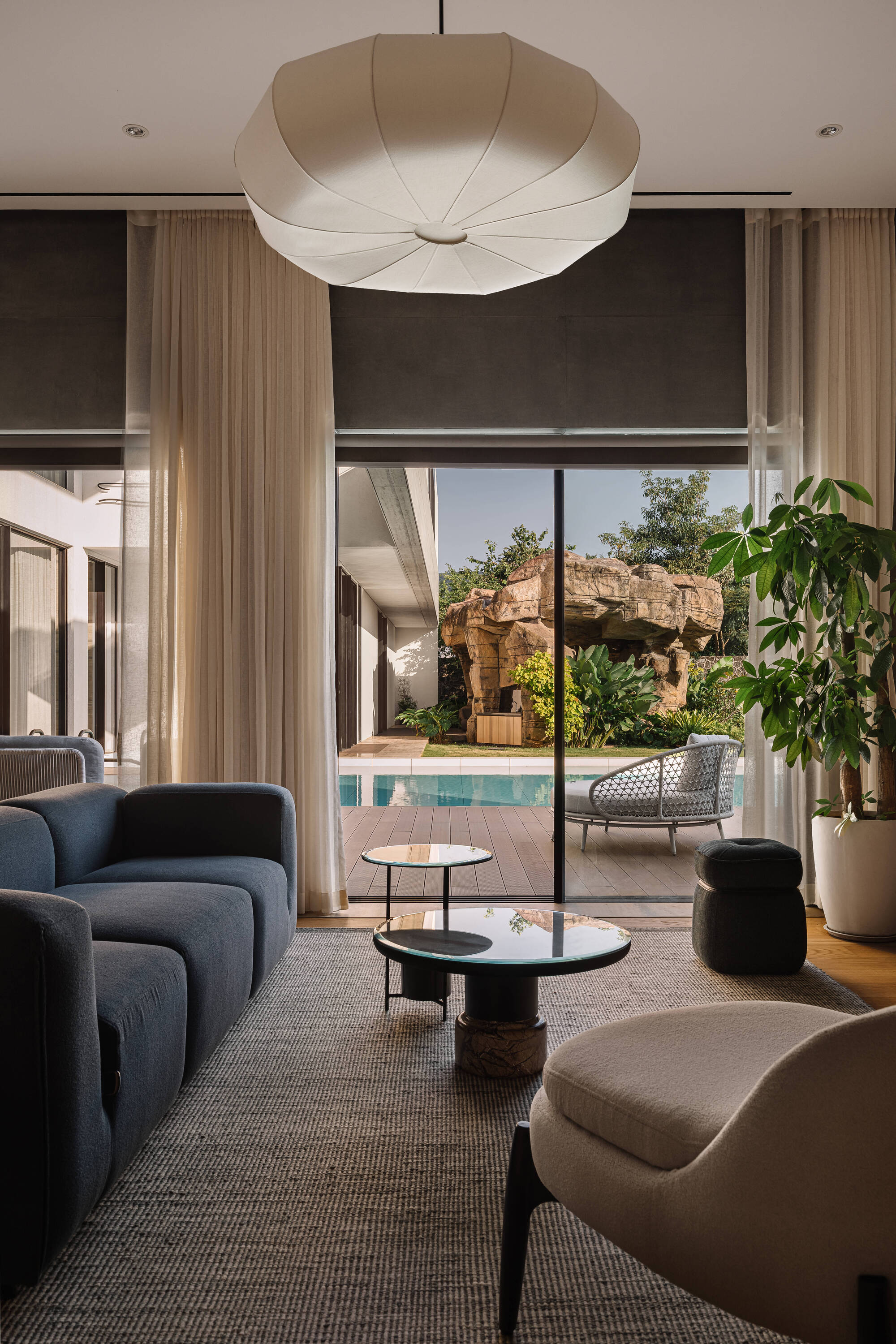 Living room view through sheer curtains and glass doors to the pool and rock feature, with a fabric pendant lamp overhead