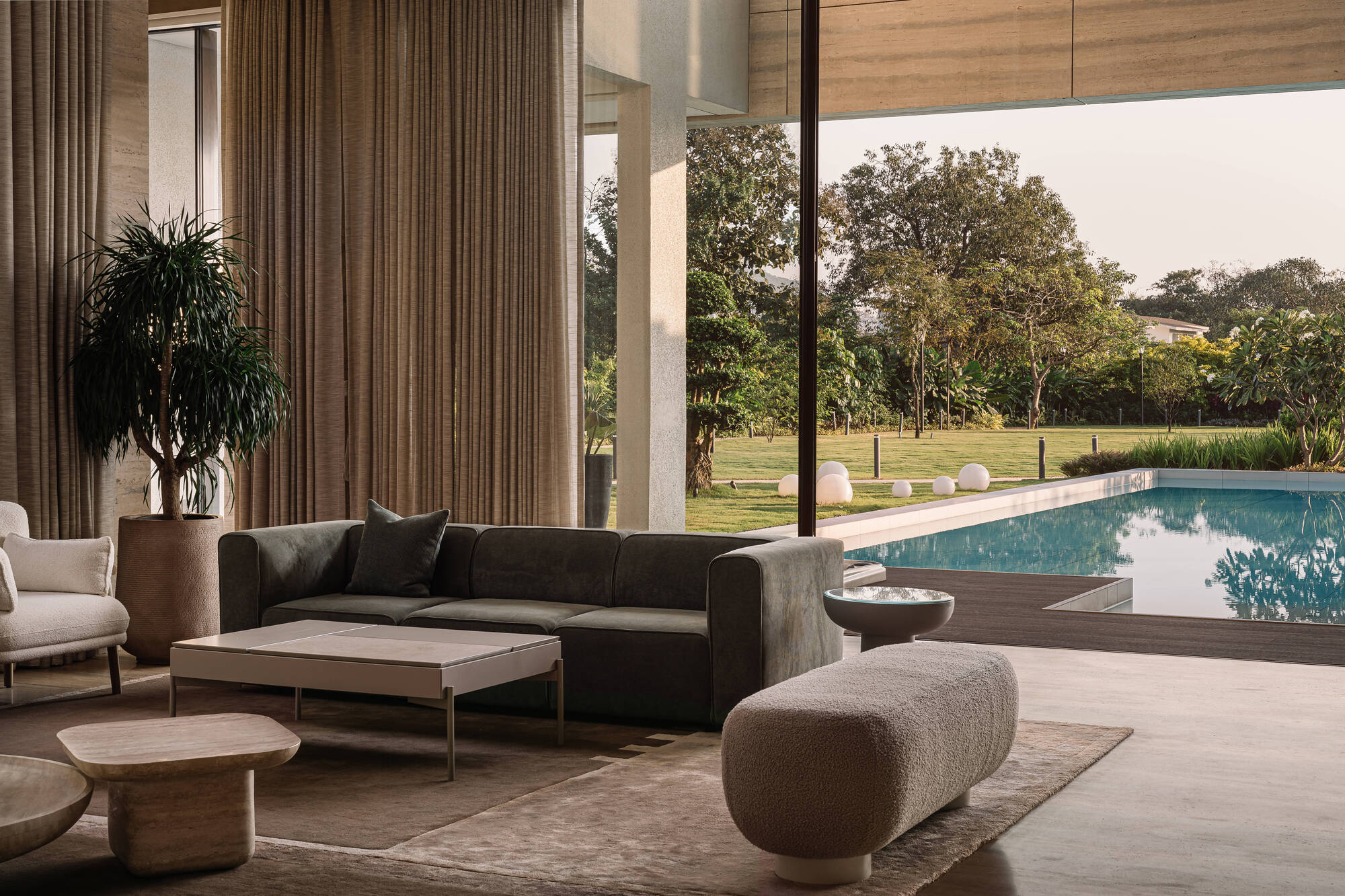 Living area with grey sofa, bouchle bench and sheer curtains opening onto the pool and a lawn of trees in afternoon light