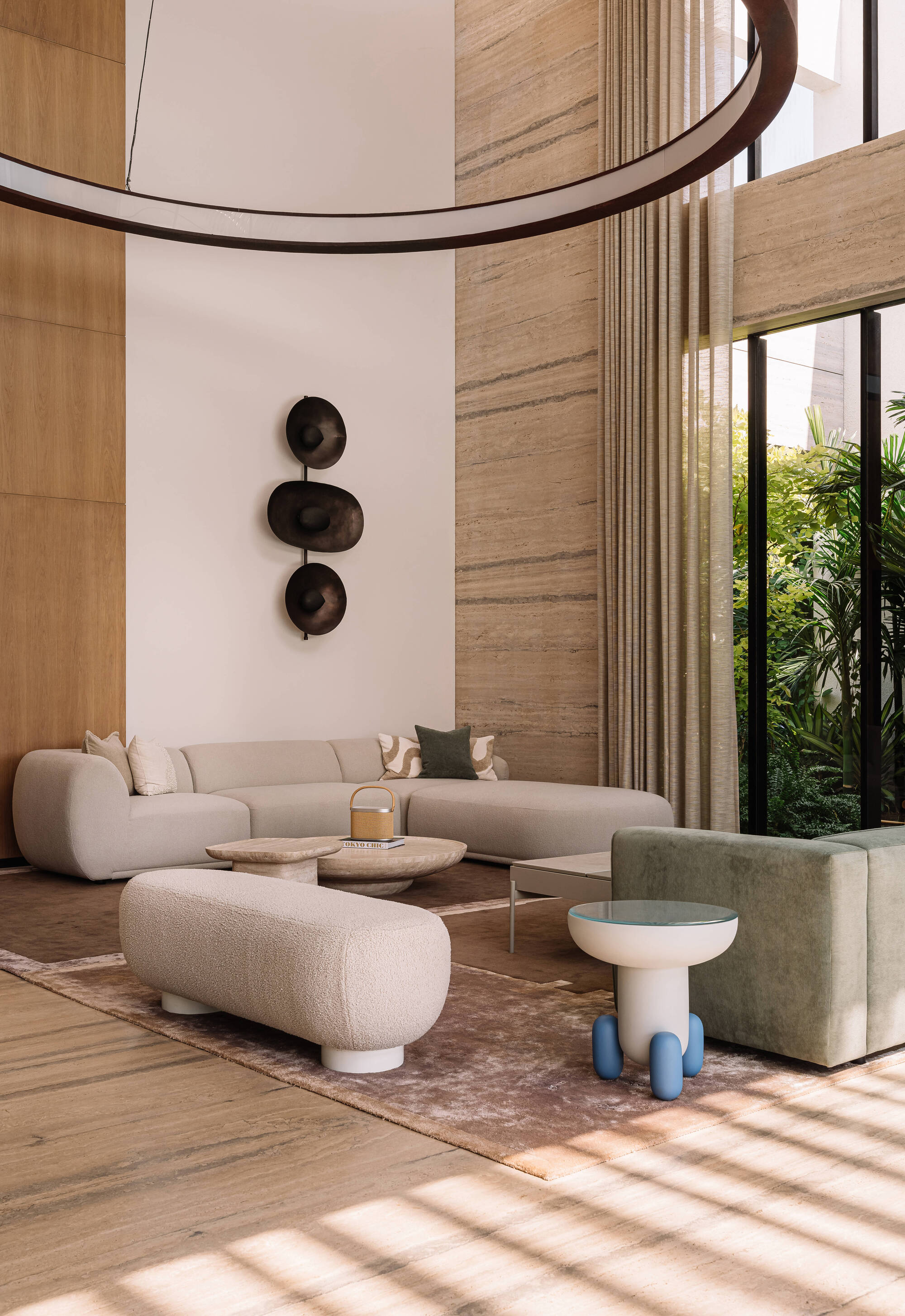 Double-height living room with curved cream sofa, travertine walls and a circular pendant, striped sunlight falling across the floor