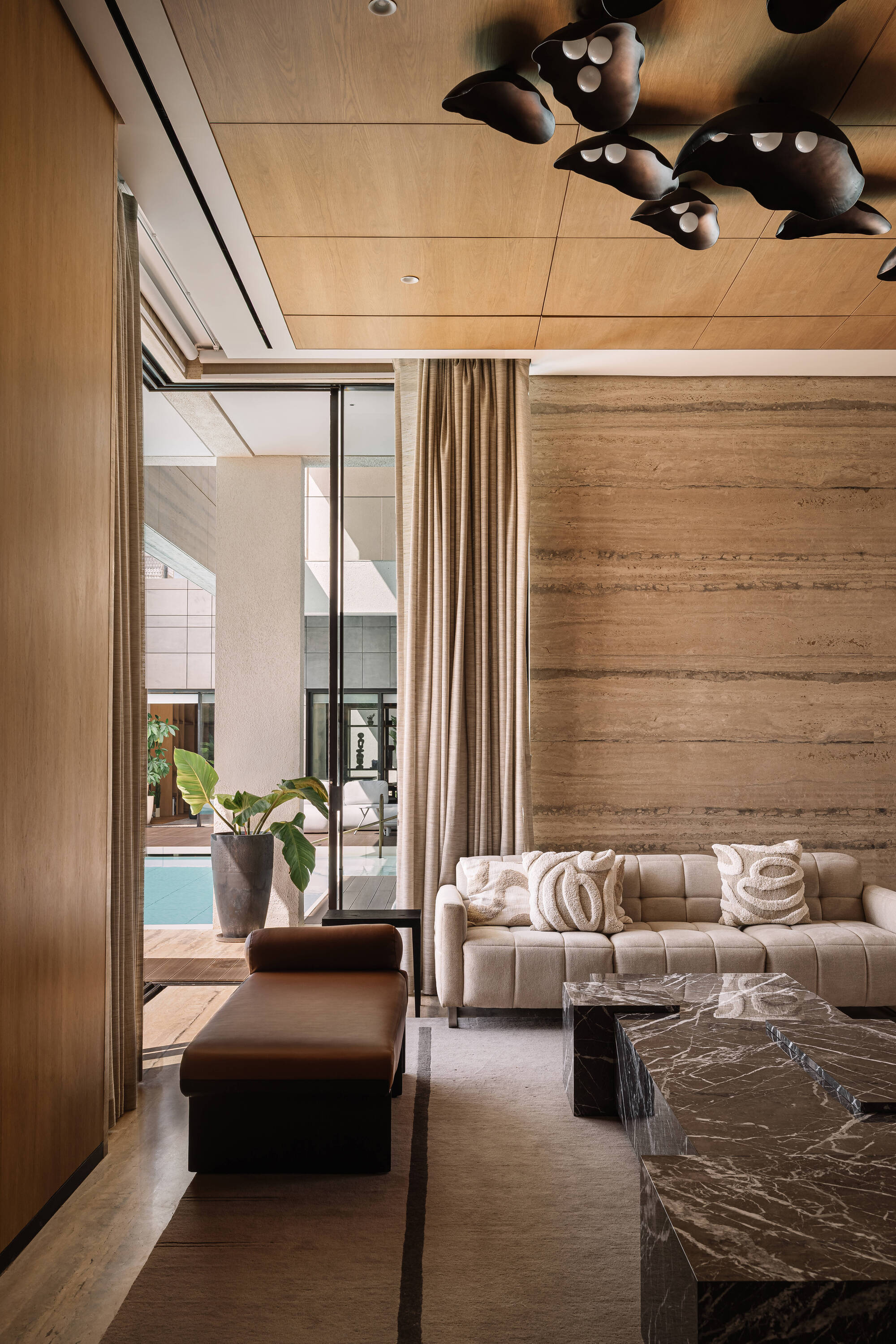 Lounge with leather bench, marble tables and travertine wall, opening through glass doors to a potted plant and pool in soft daylight