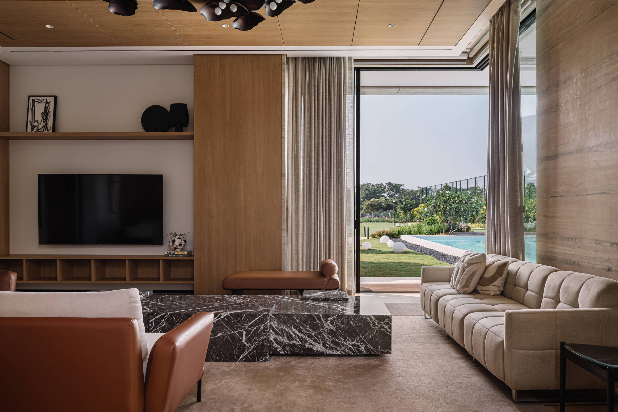 Living room with built-in shelving, leather armchair and marble table, sliding glass doors open to a lawn and pool under clear sky
