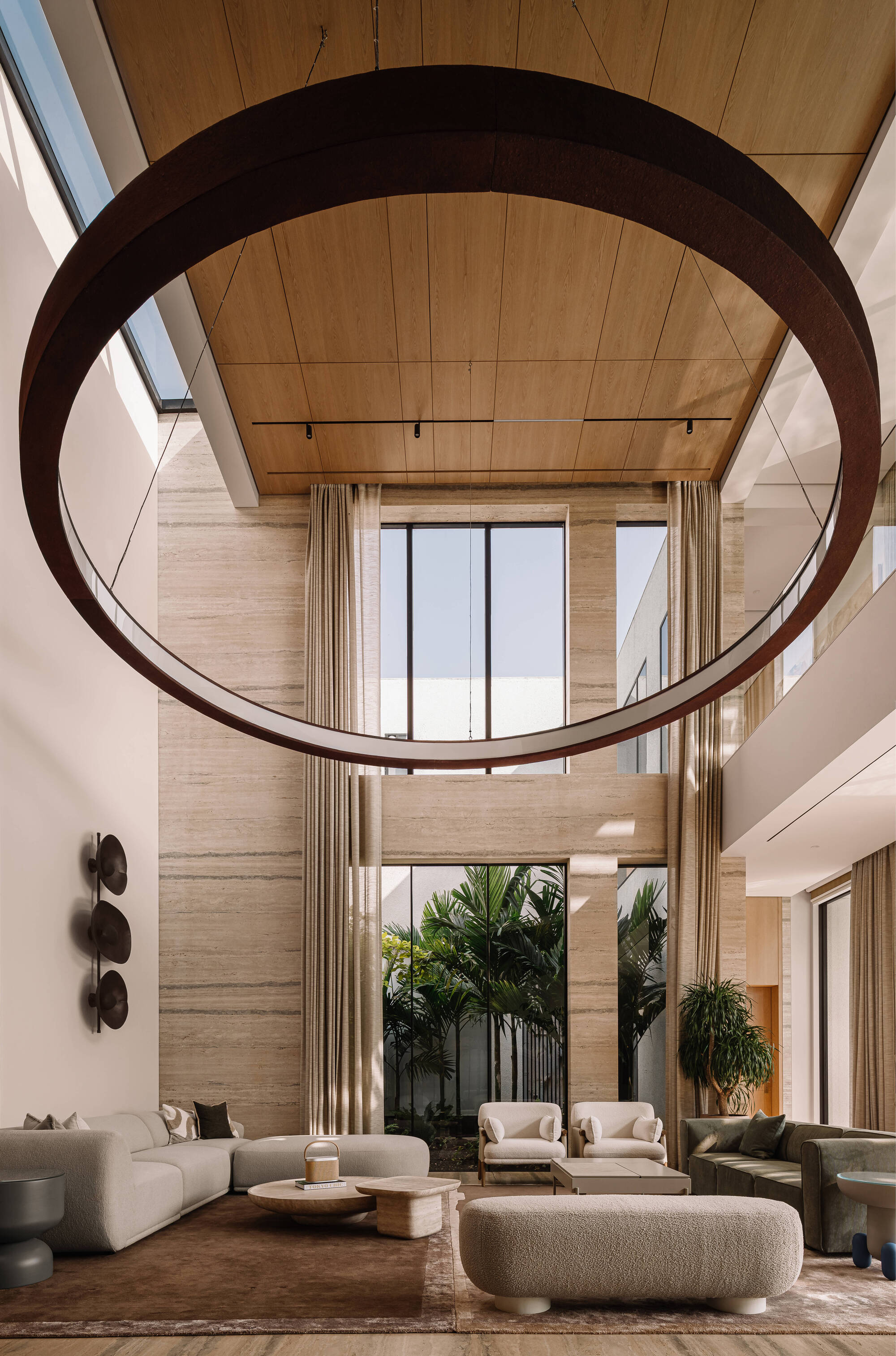 Double-height living room with a weathered steel ring pendant hanging below a timber ceiling, travertine walls and a palm courtyard beyond