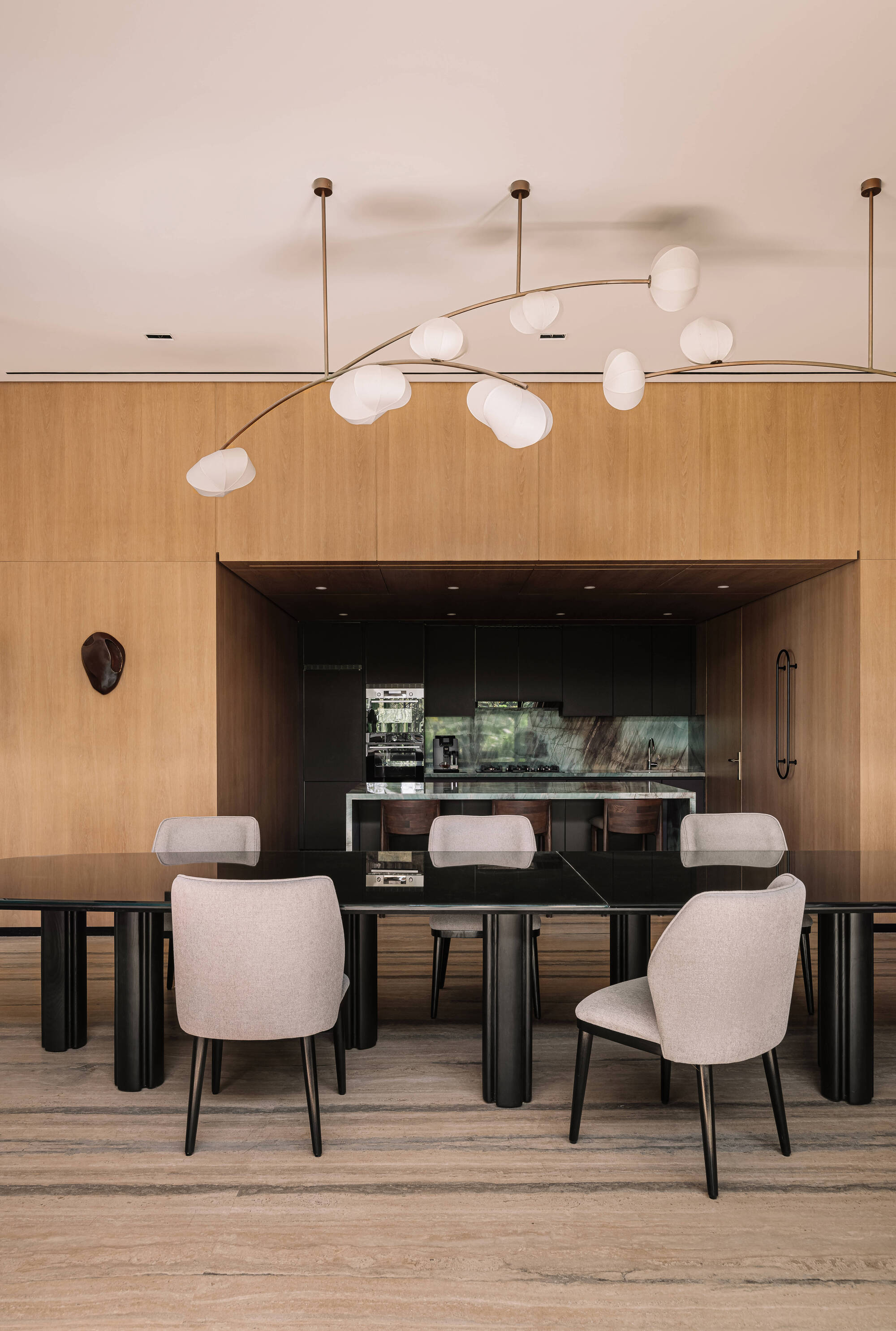 Dining area with a black stone table, upholstered chairs and a branching globe pendant in front of a timber-clad kitchen alcove