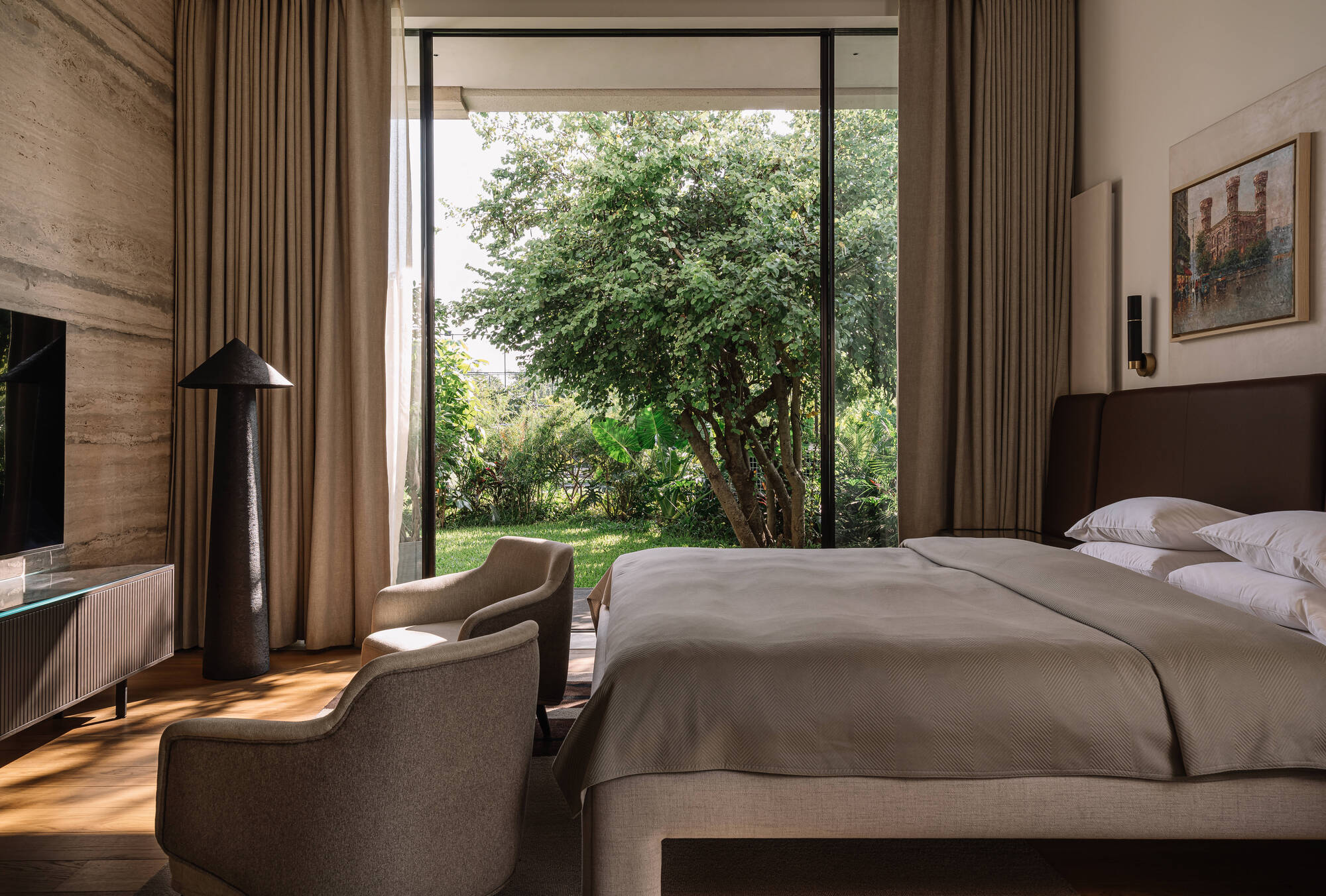 Bedroom with floor-to-ceiling glazing framing a leafy tree and lawn, with linen curtains and a pair of armchairs in afternoon light
