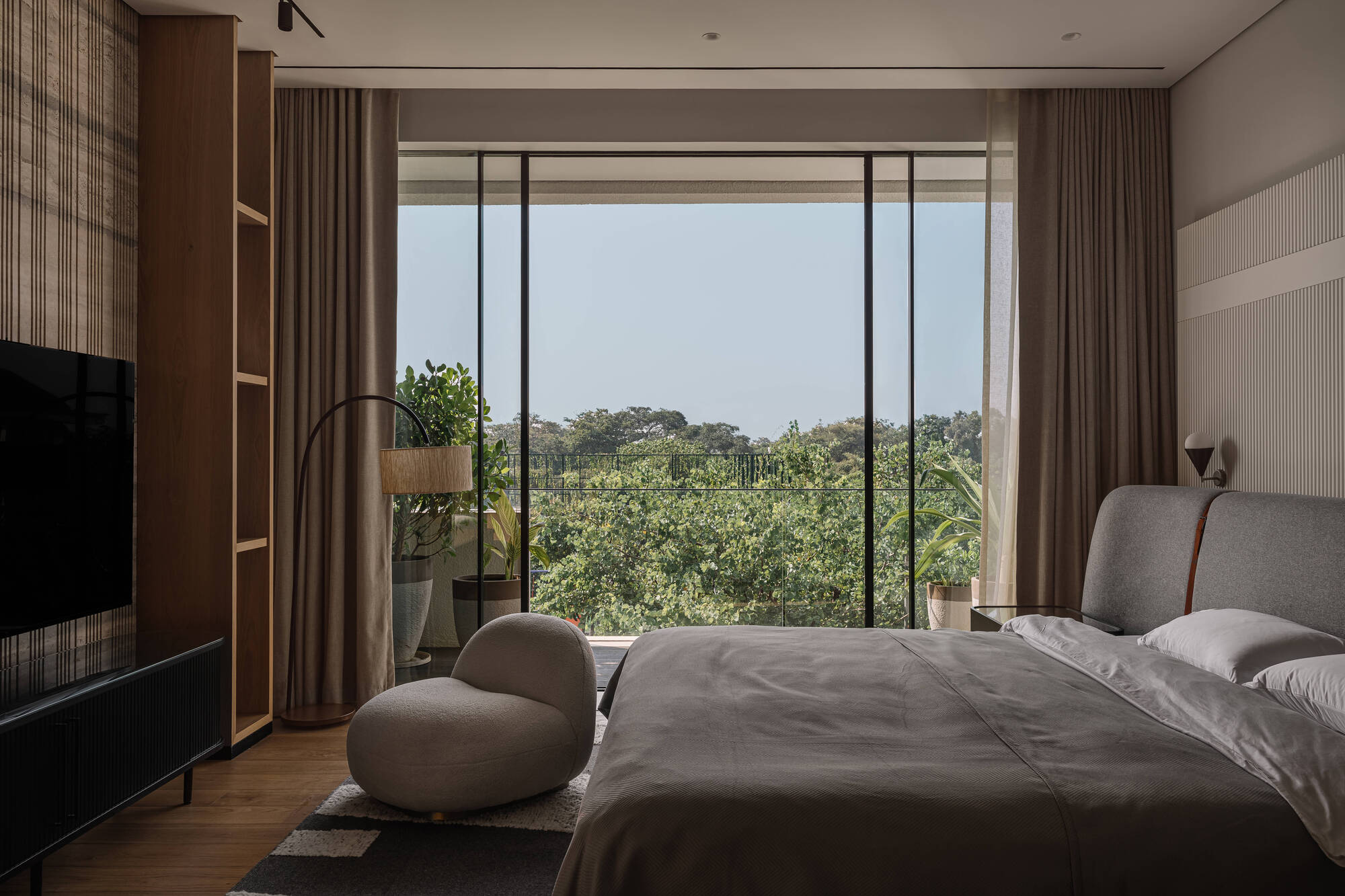 Bedroom with a grey upholstered bed and round pouf facing full-height glass that opens onto a canopy of treetops under clear sky