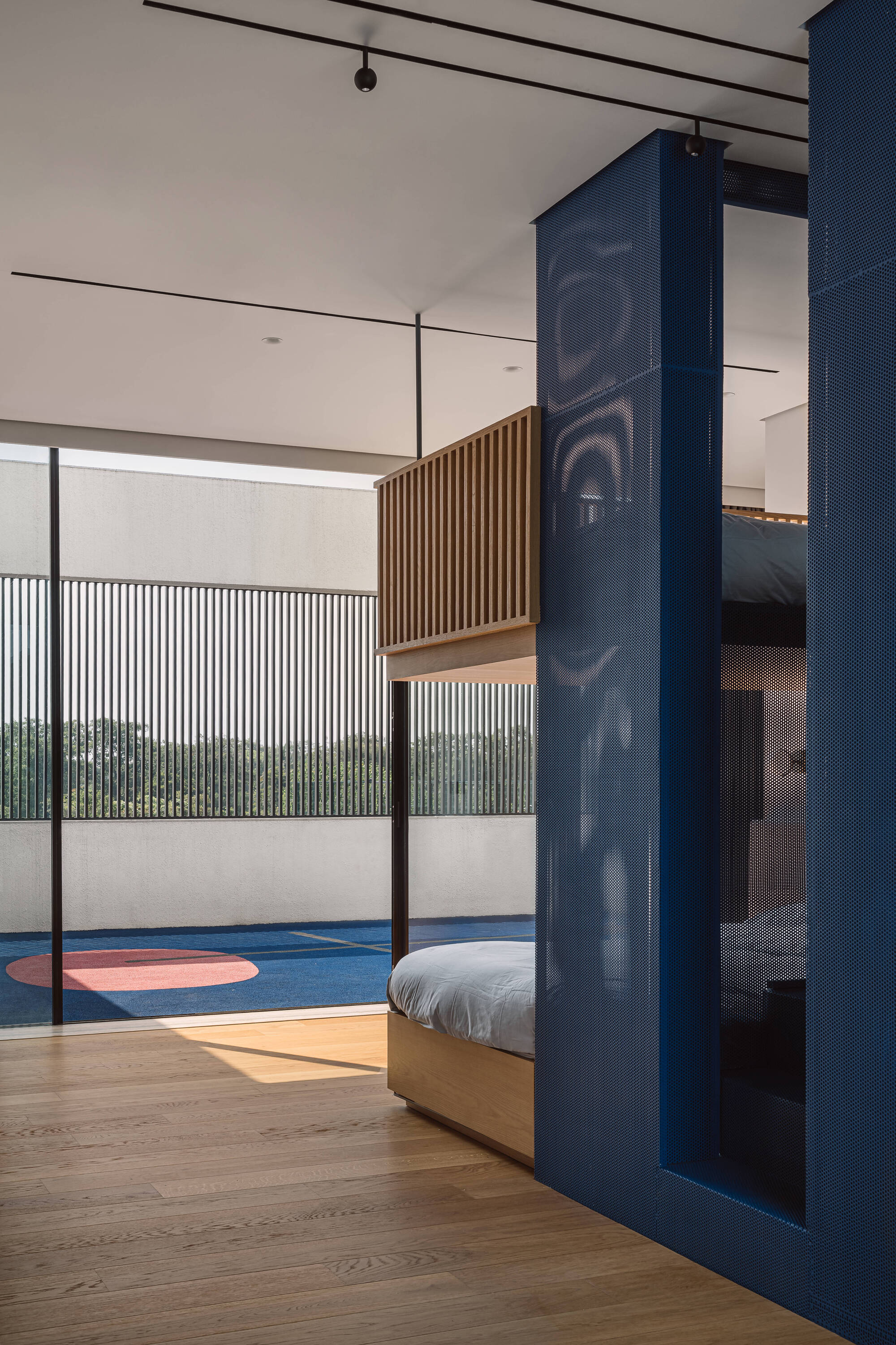 Blue perforated metal volume enclosing a bunk bed, beside a glazed wall opening onto a terrace with a painted court circle