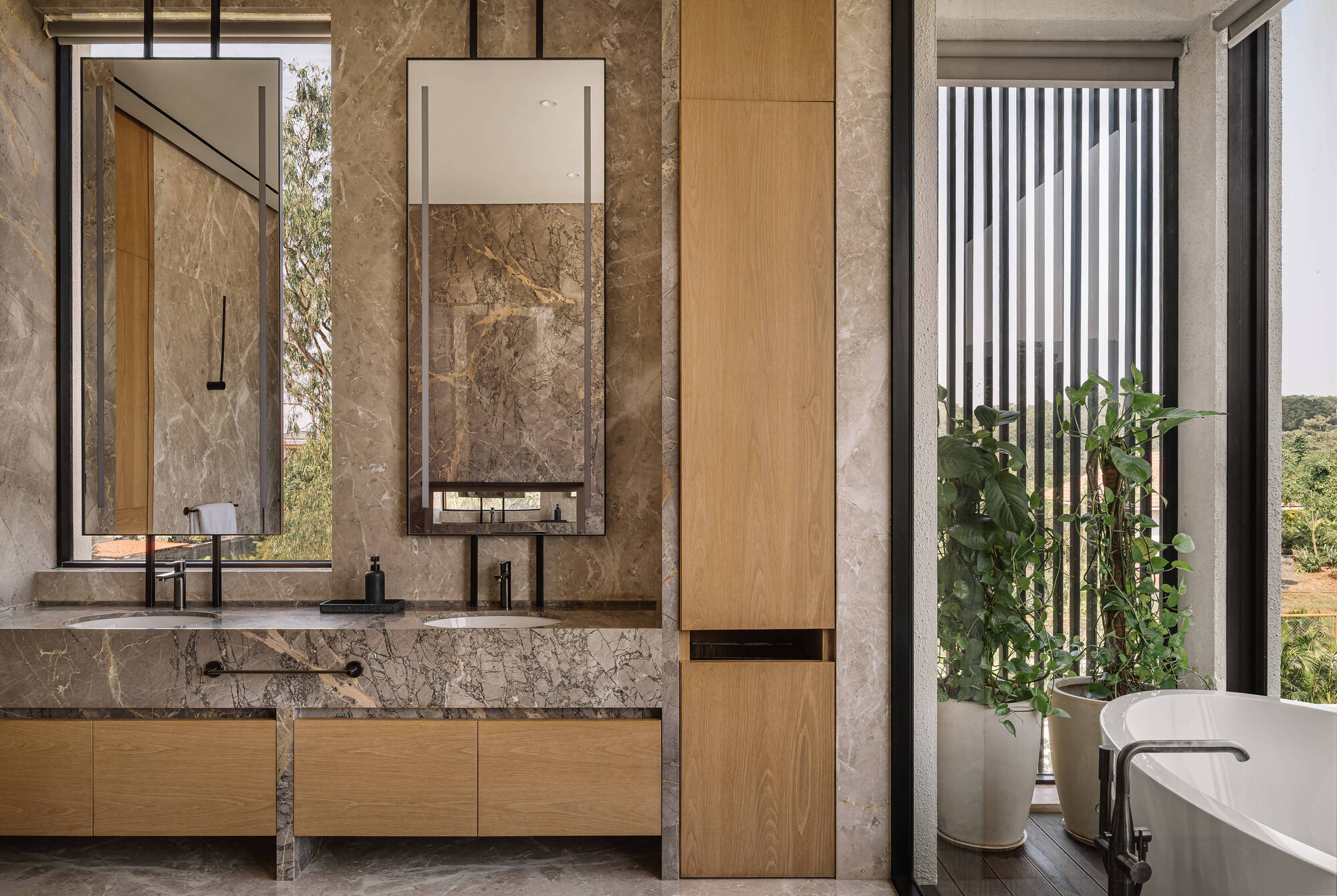 Bathroom with a marble double vanity, hanging mirrors and oak cabinetry beside a freestanding tub and potted plants by the window