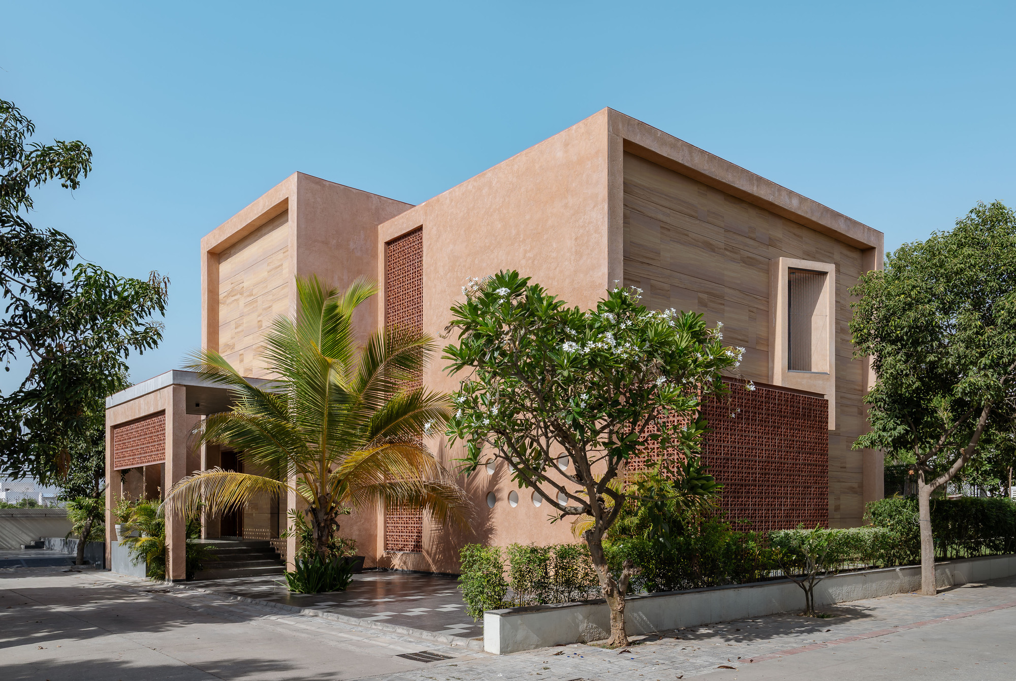 Corner view of the blush plastered volumes with stone cladding and brick lattice screens, framed by a palm and flowering frangipani in clear sun