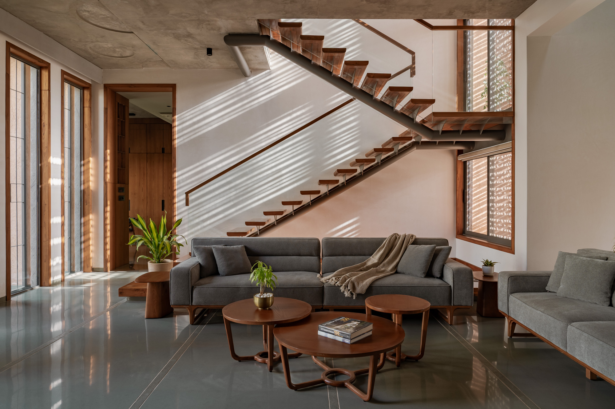 Living room with a floating timber-tread staircase, grey sofa and round wood tables, striped sunlight cast across the white wall