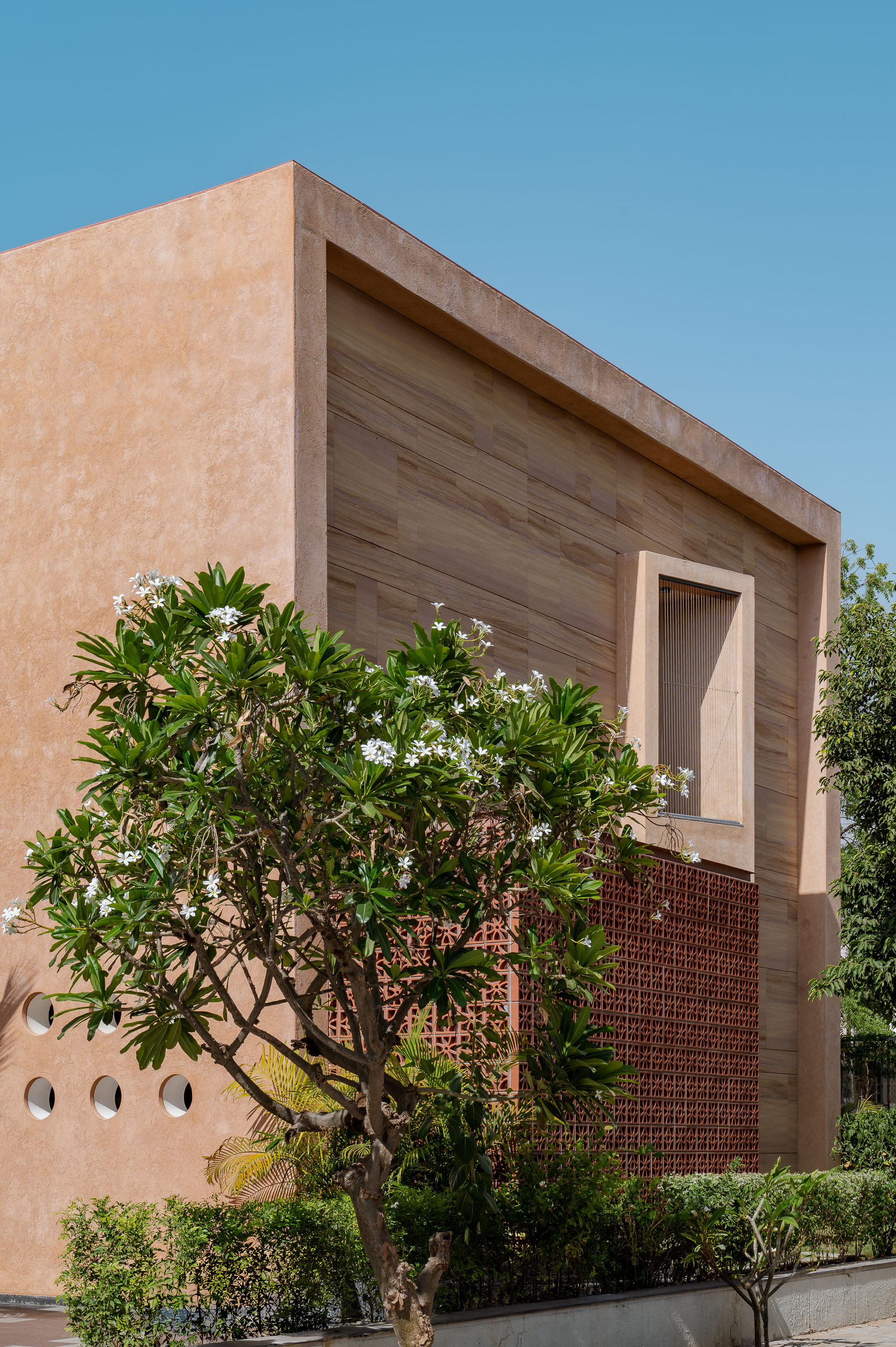 Stone-clad upper volume with a recessed window above a terracotta lattice screen, a flowering frangipani tree in the foreground