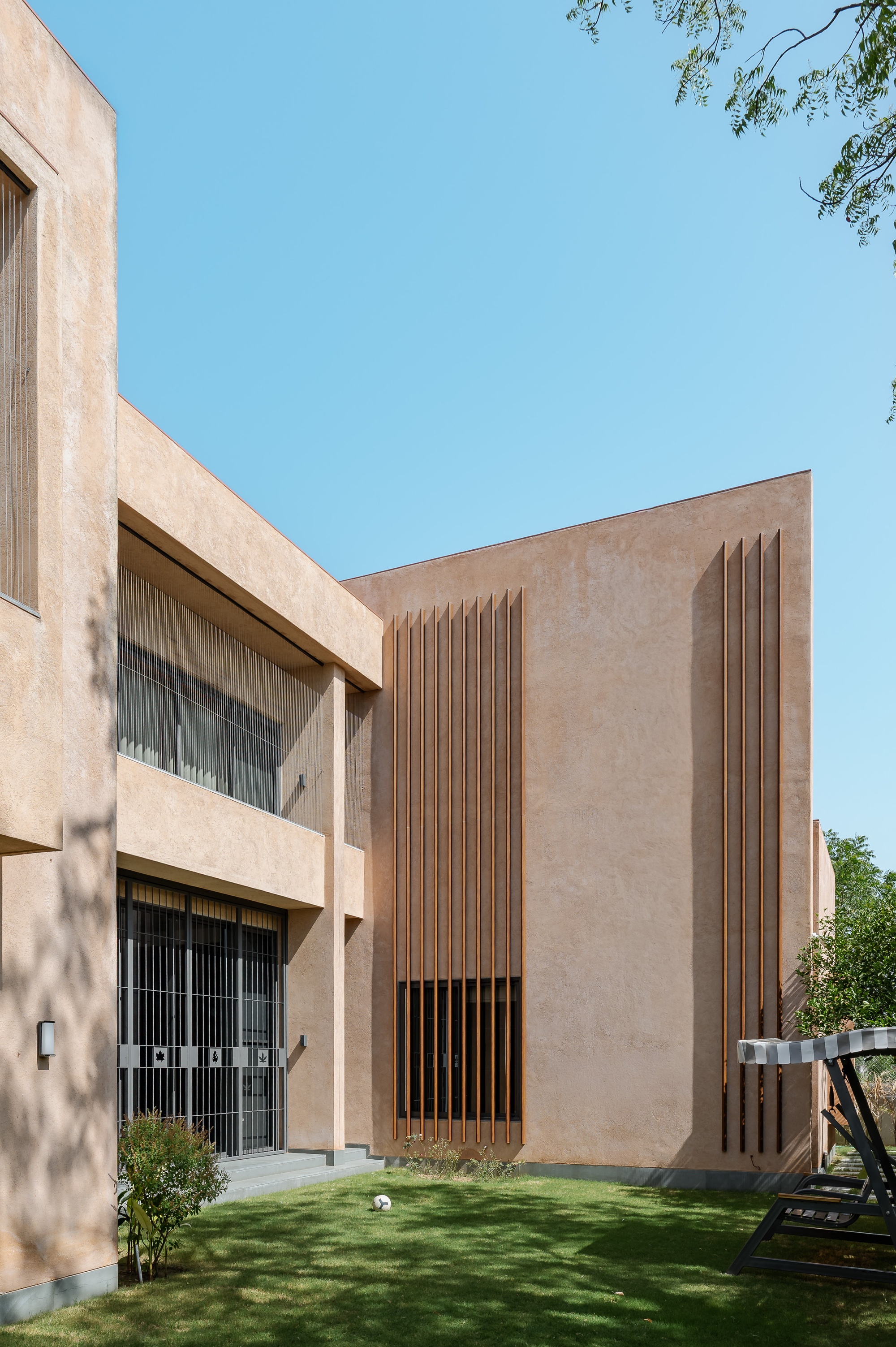 Plastered ochre facade with tall vertical timber fins and a barred ground-floor window, seen across a lawn under clear blue sky