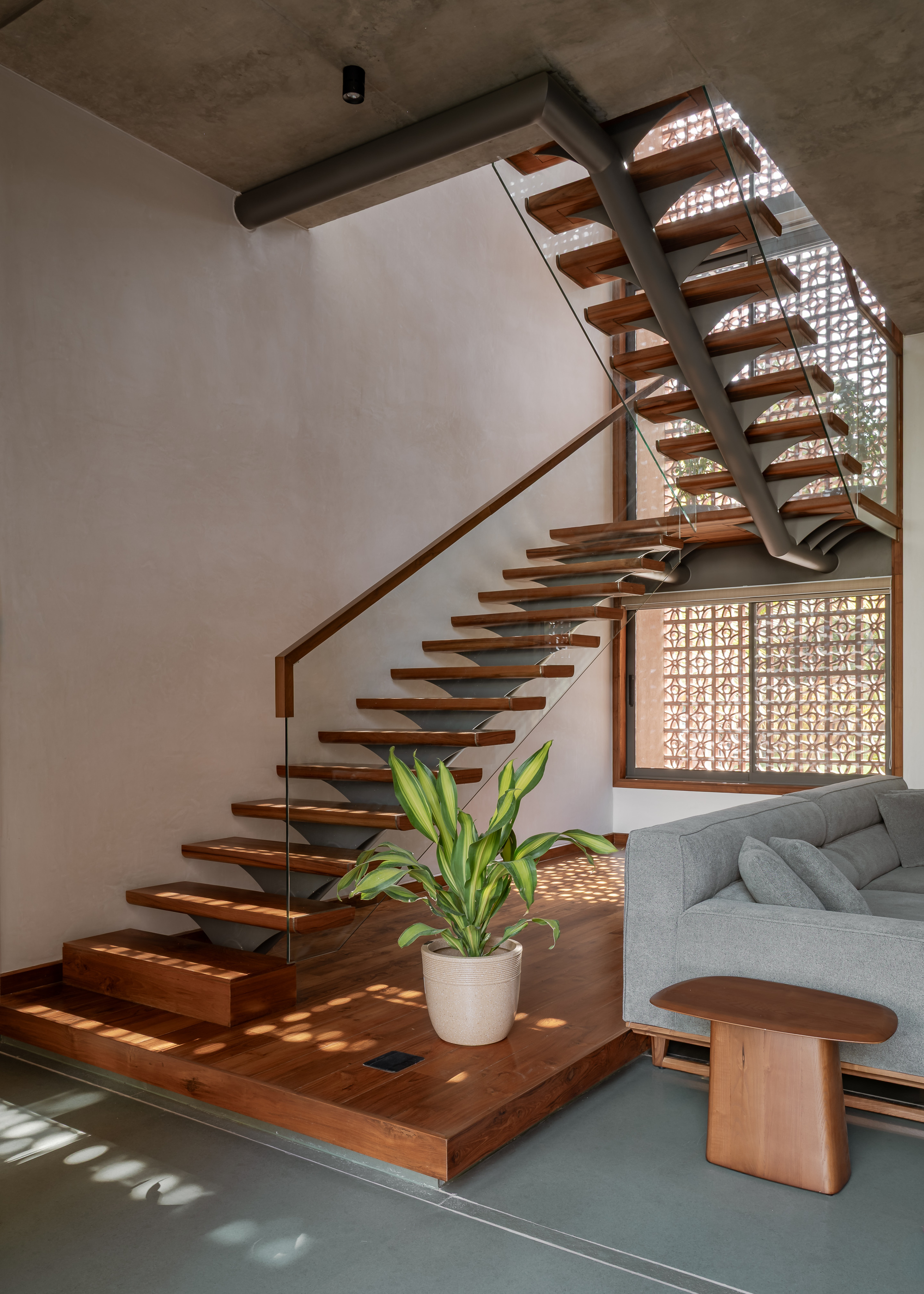Open-riser timber staircase with glass balustrade beside a perforated screen window, casting dappled sunlight patterns across the wooden platform
