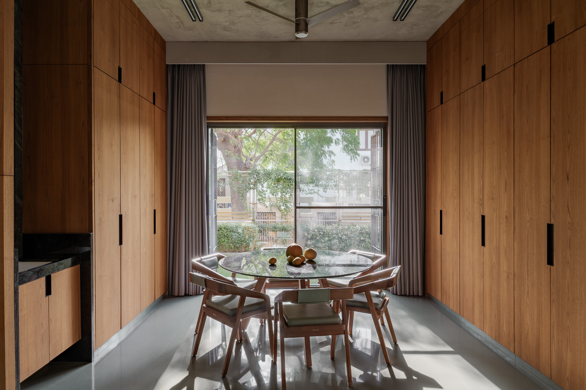 Dining nook with a round glass table and wooden chairs between floor-to-ceiling timber cabinets, facing a window onto a leafy tree