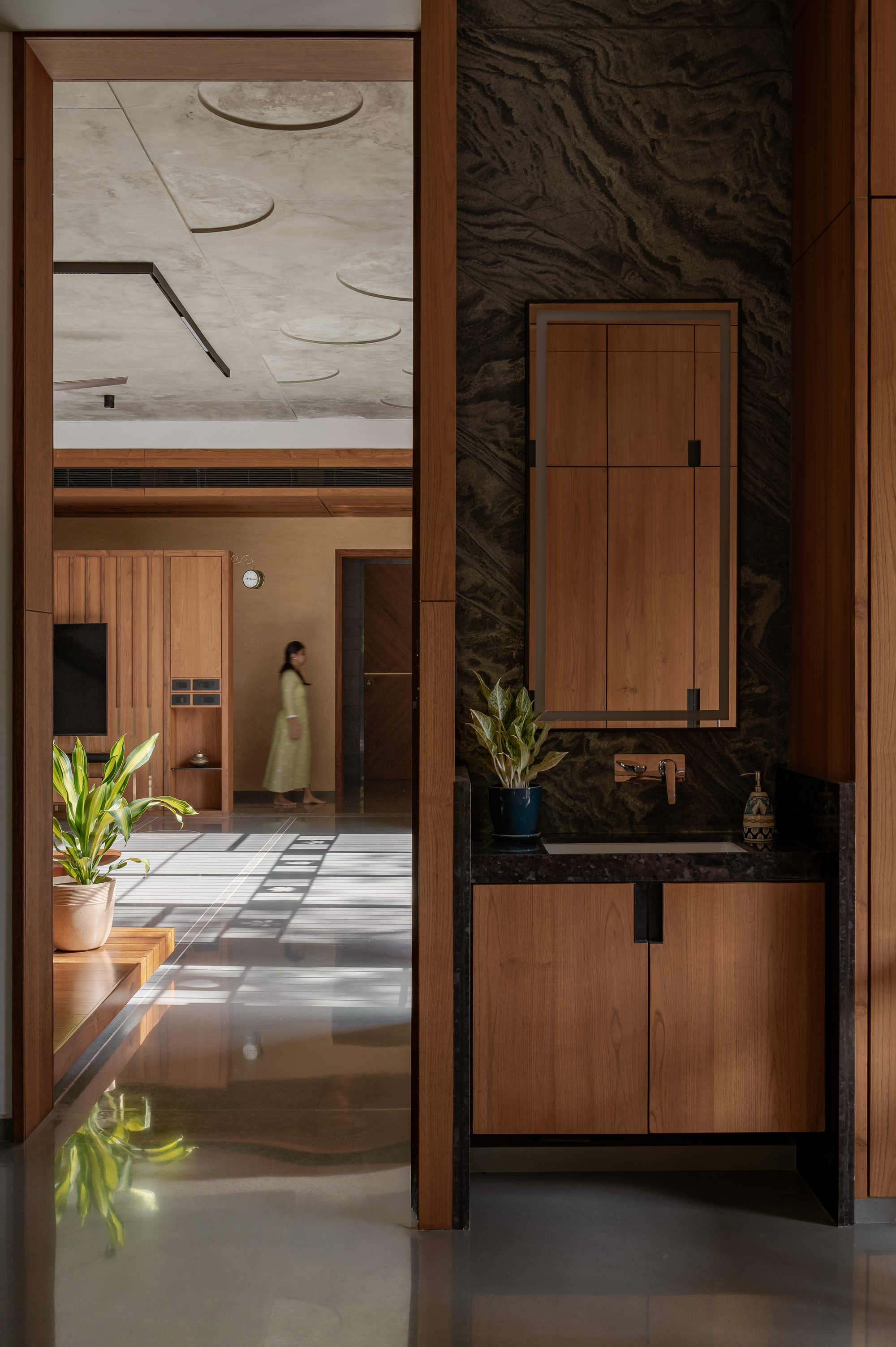 Wash basin alcove with dark veined stone backdrop and timber cabinets, while a woman walks through the sunlit living room beyond
