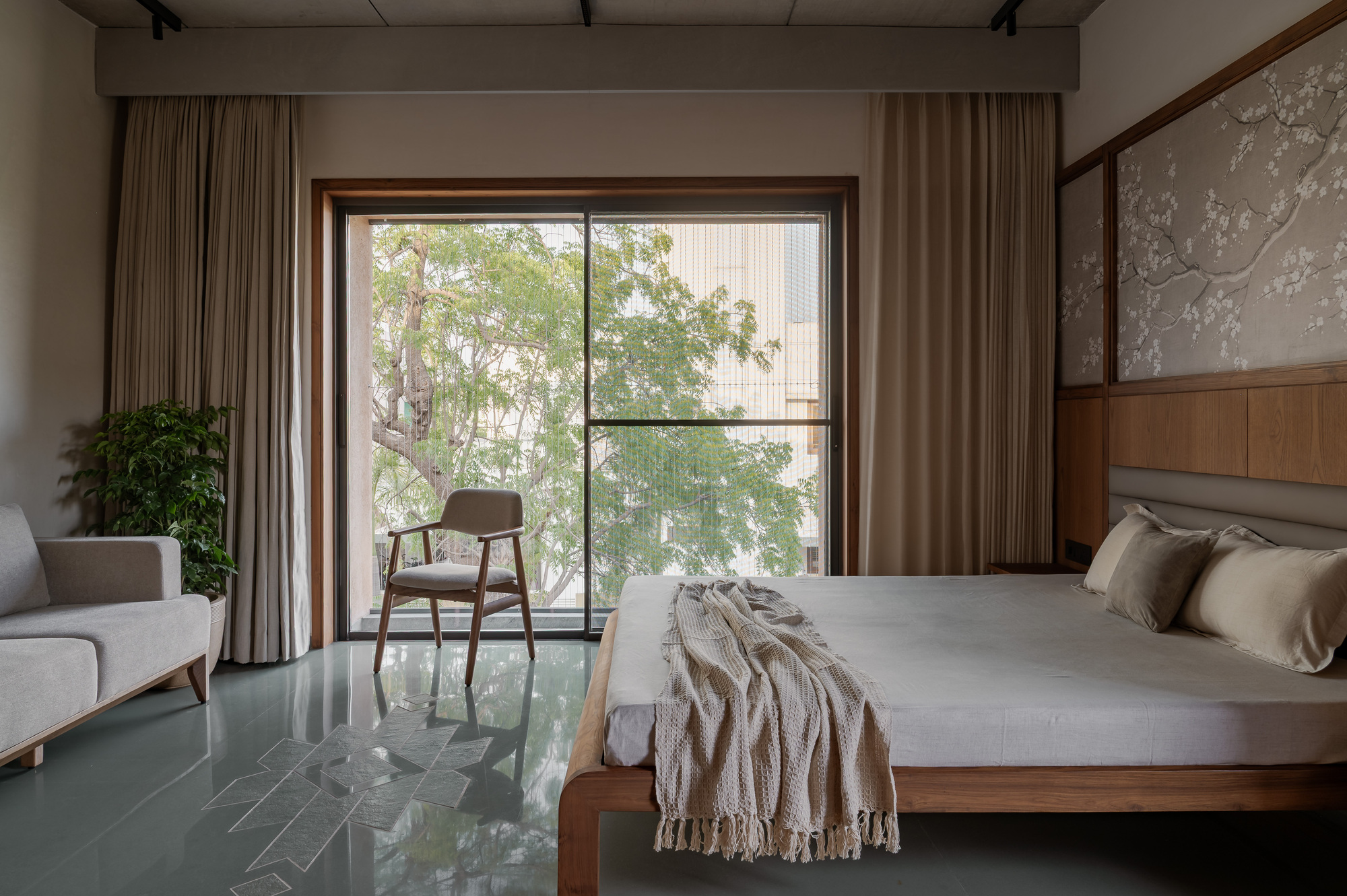Bedroom with a low timber bed, floral wall panel and sliding glass door framing a leafy tree, with a chair in daylight