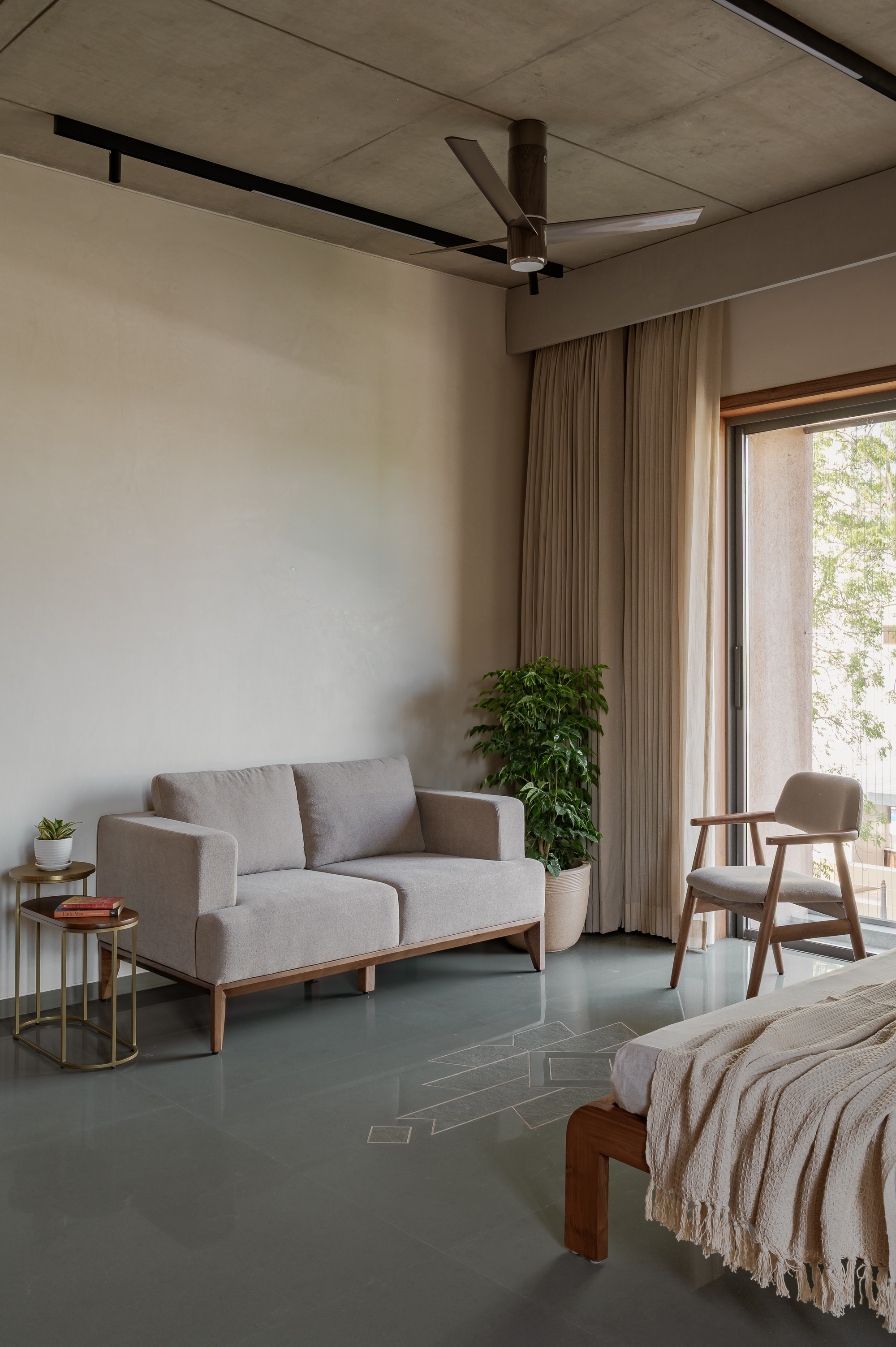 Bedroom corner with a grey fabric loveseat, potted plant and ceiling fan under a concrete soffit, daylight entering through a sliding glass door