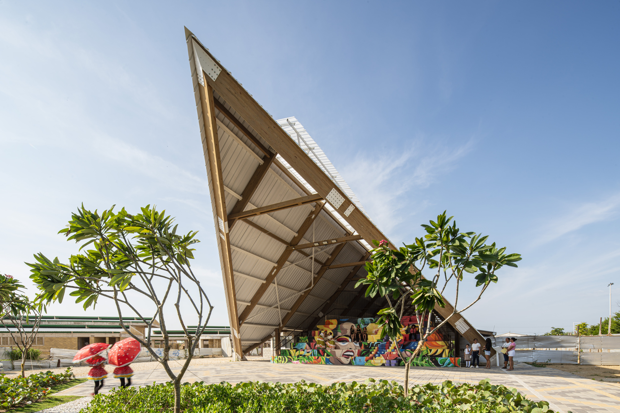Tilted timber-framed roof plane rising over a colorful mural wall, with two figures carrying red umbrellas past frangipani trees