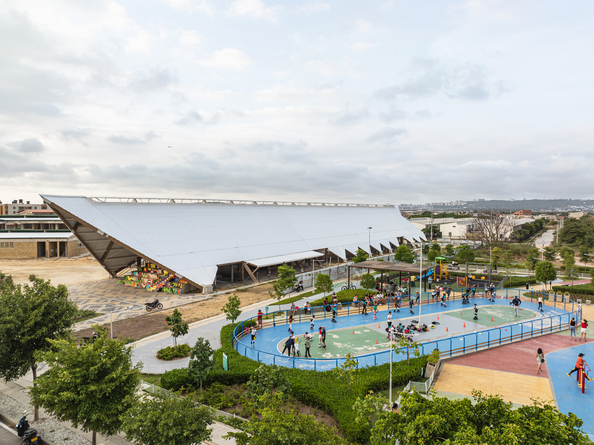 Aerial view of the long white pitched roof beside a blue skating rink with children playing under an overcast sky