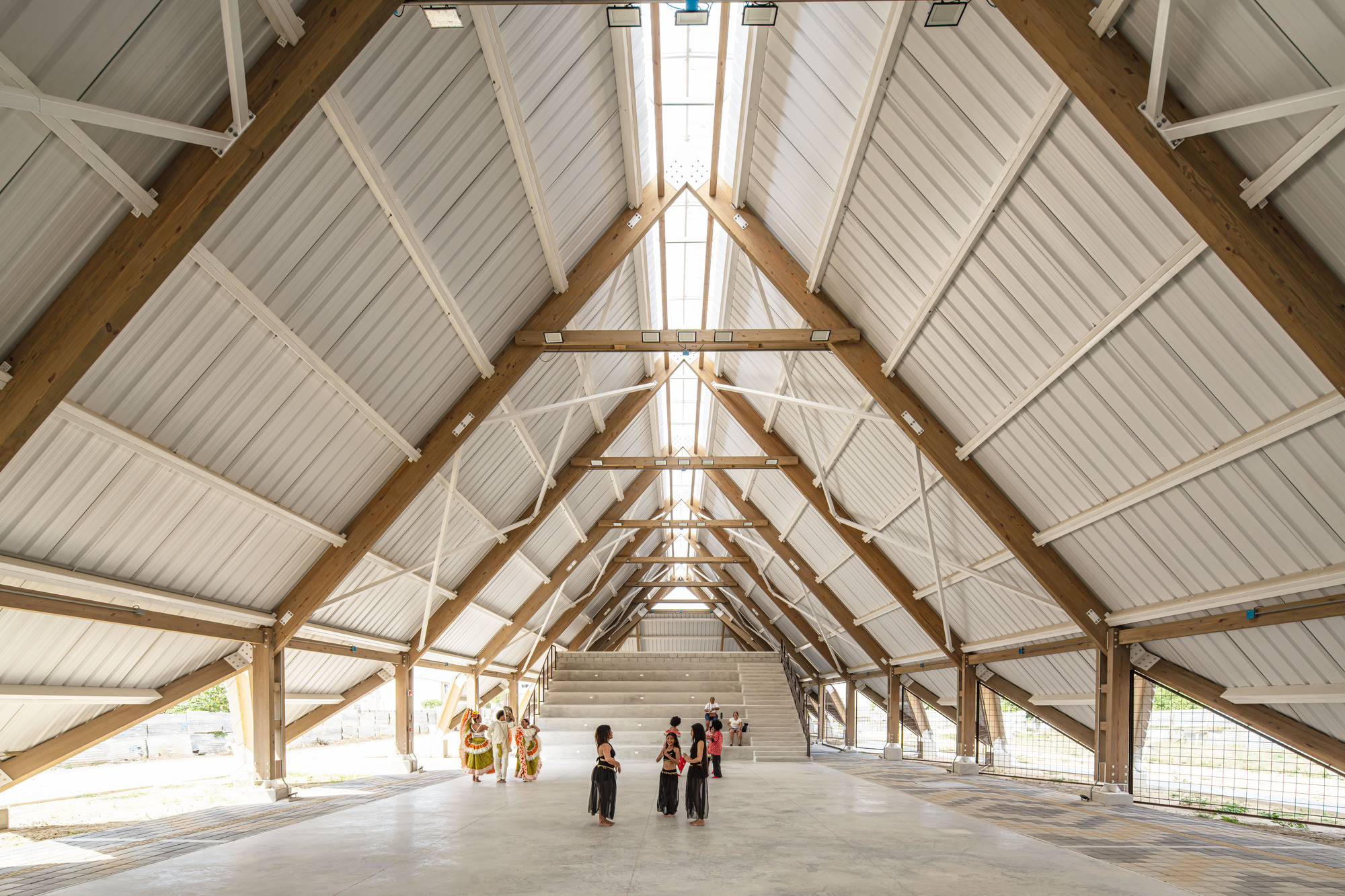 Interior of the open hall with glulam timber trusses, corrugated roof panels and a group of dancers on the concrete floor