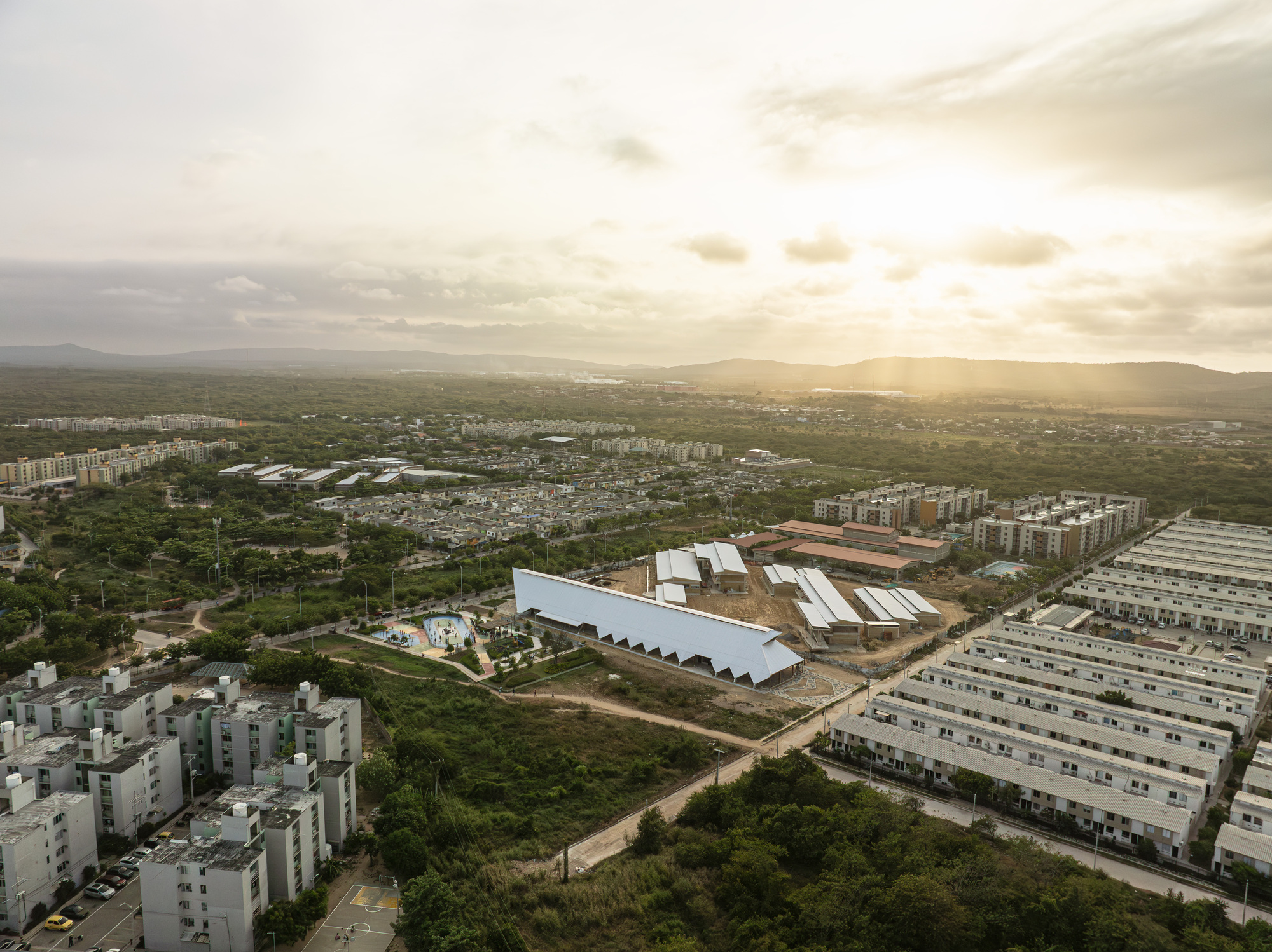Distant aerial view of the white-roofed building among housing blocks and green scrub, with hills under low evening sun