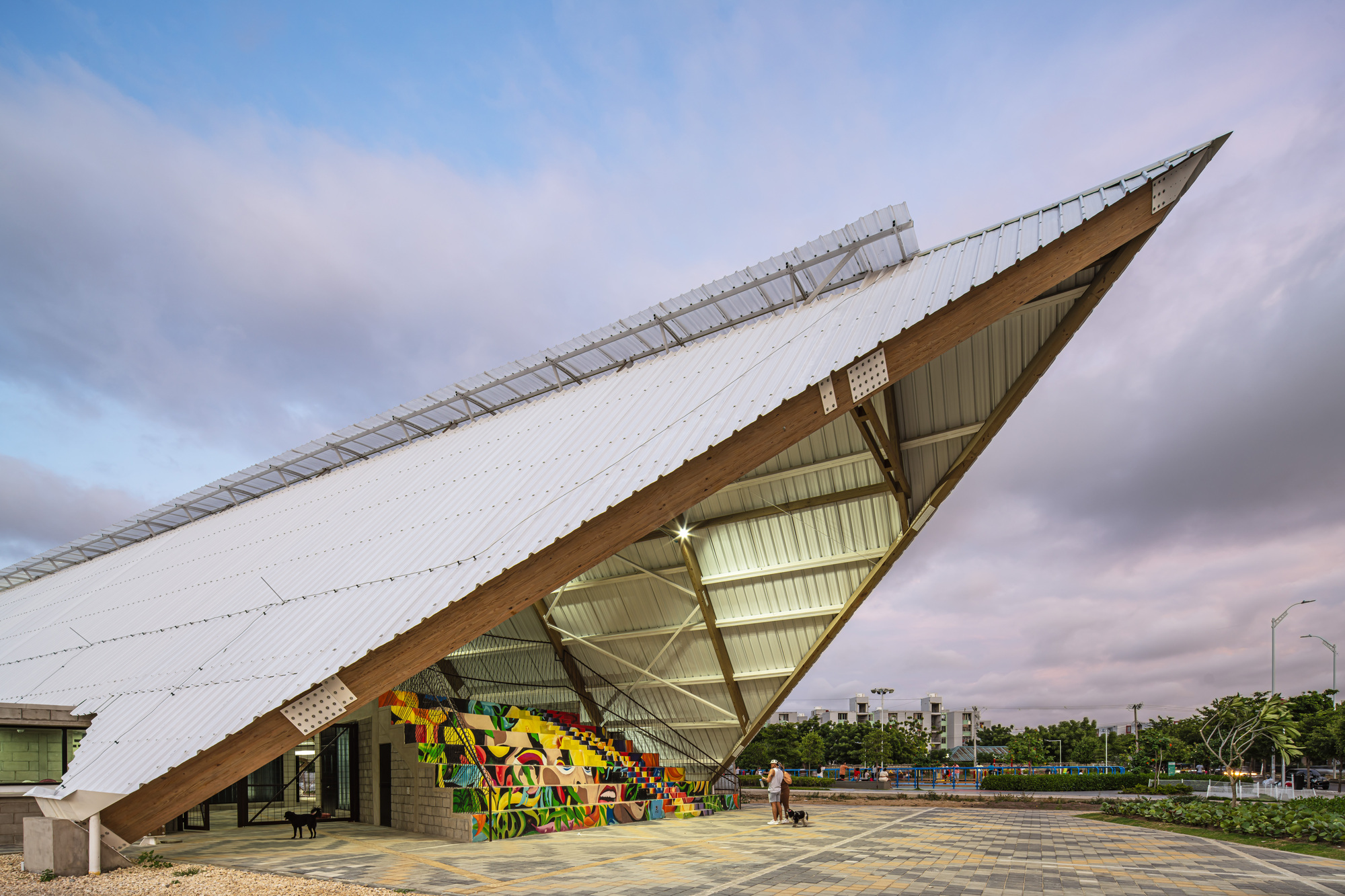Corner of the sloping white roof lifted on timber beams above a graffiti-painted stepped seating wall at dusk